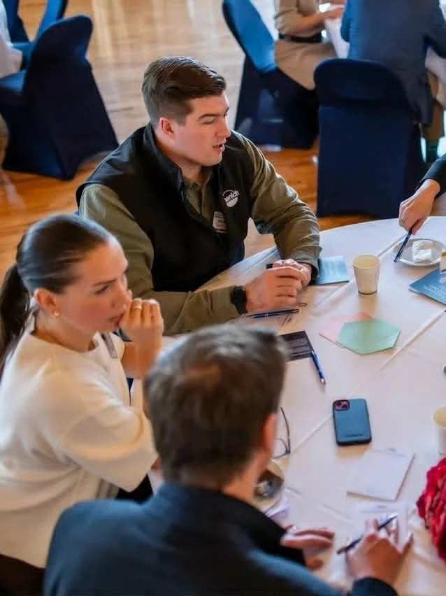 People sitting around a table at a meeting or event, engaged in discussion with notes, pens, and drinks on the table.