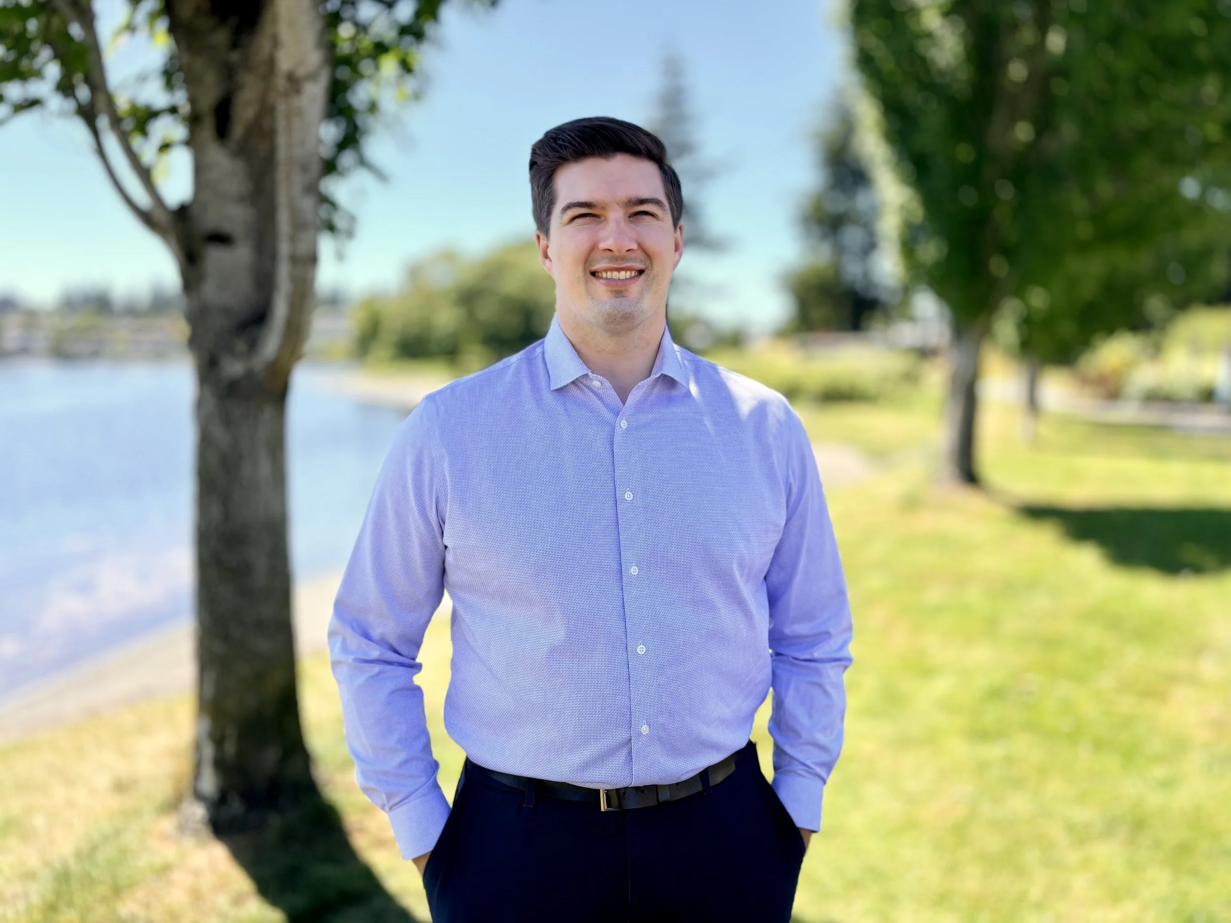 A man in a light blue button-up shirt with dark pants standing outdoors in front of trees and a body of water, smiling.