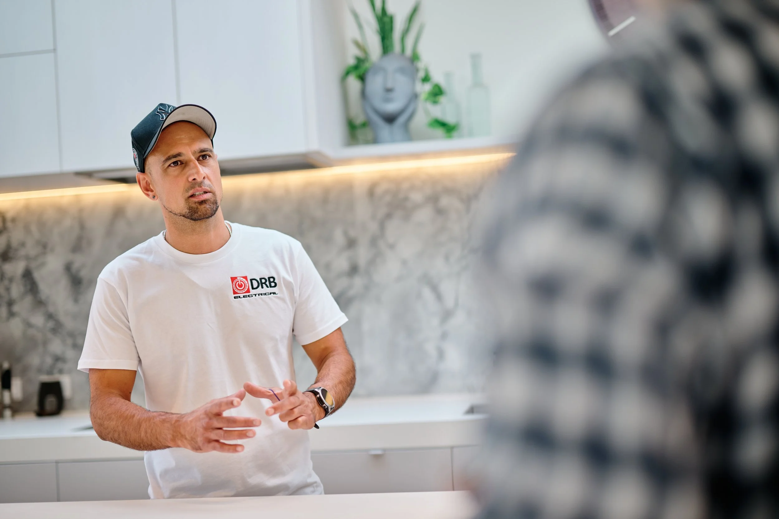 A man wearing a white t-shirt with a logo, a black cap, and a watch, is standing indoors in front of a white kitchen with modern decor. There is a decorative sculpture and green plants on the wall in the background.
