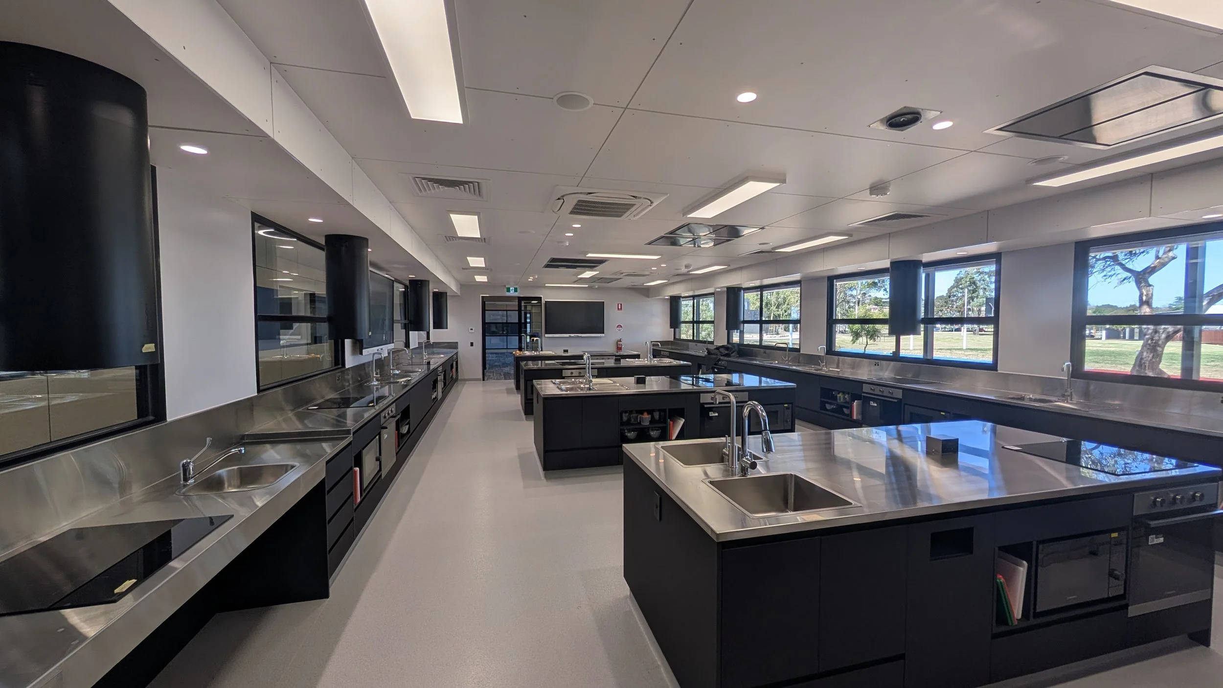 Modern school laboratory with multiple stainless steel sinks and black cabinets, large windows with outdoor view of trees, ceiling with recessed lighting and vents.