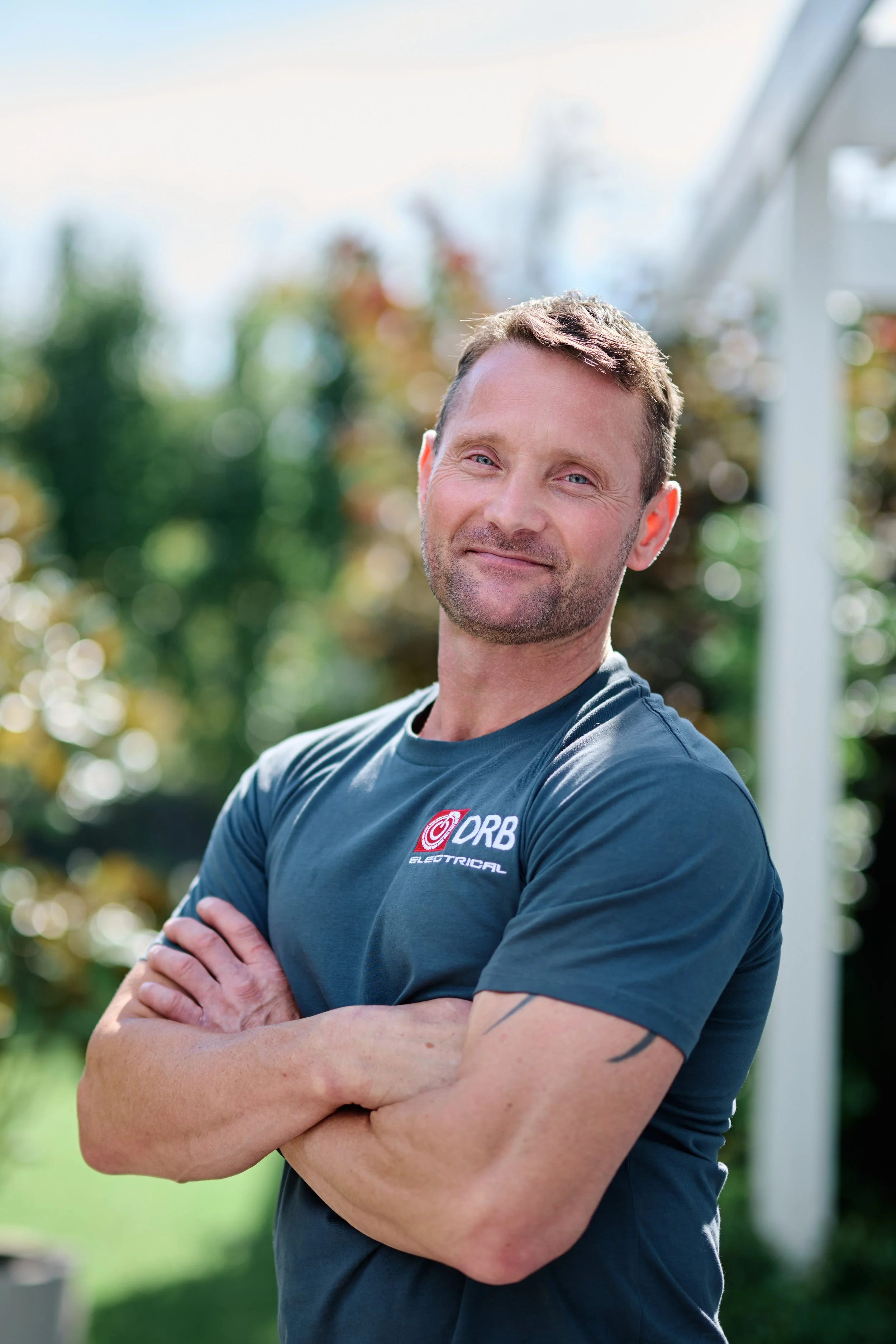 A man with crossed arms smiling outdoors with blurred trees and sky in the background.
