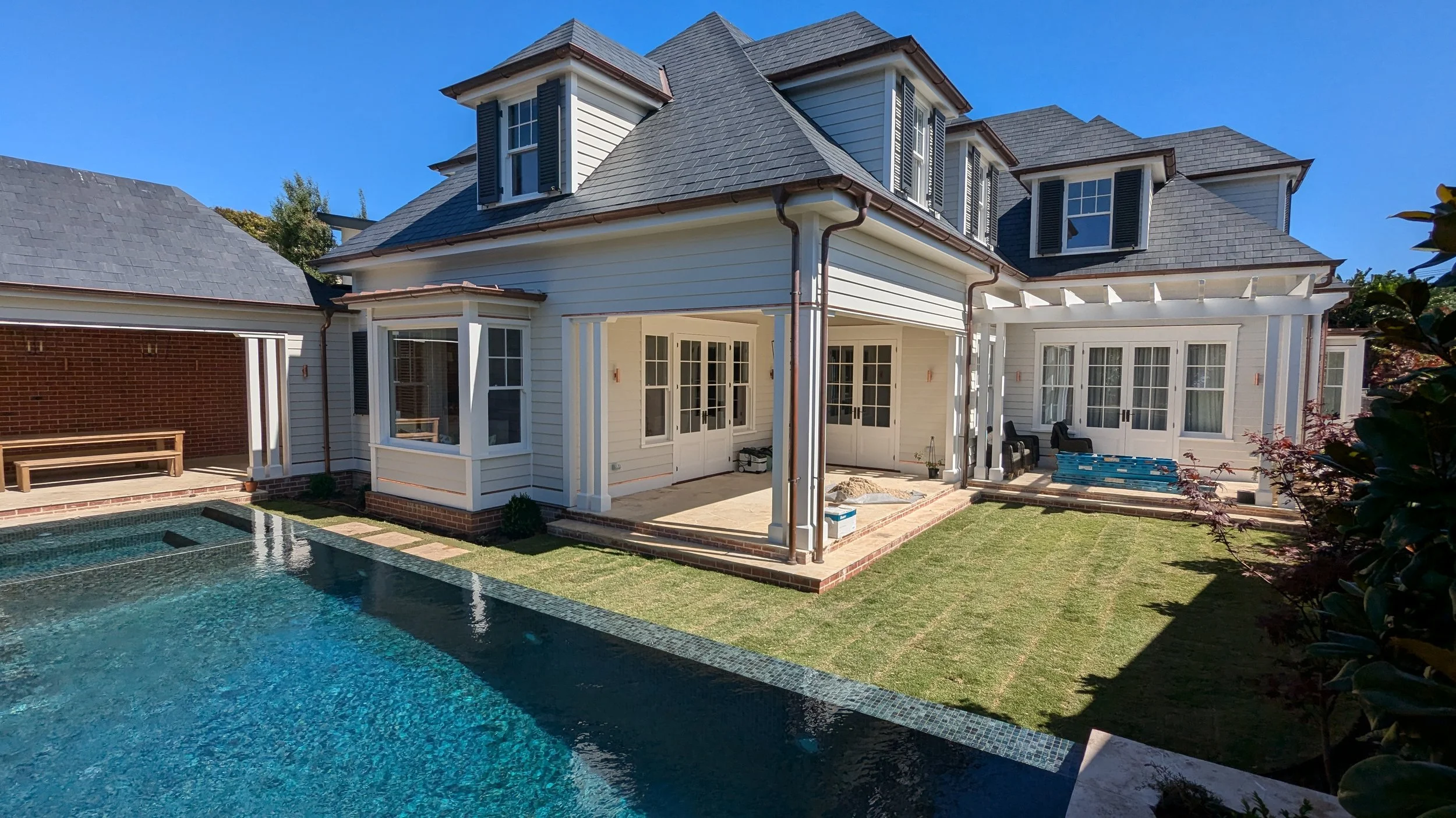 Backyard view of a white house with black shutters, multiple dormer windows, a patio, a swimming pool, and a lawn with bushes against a bright blue sky.