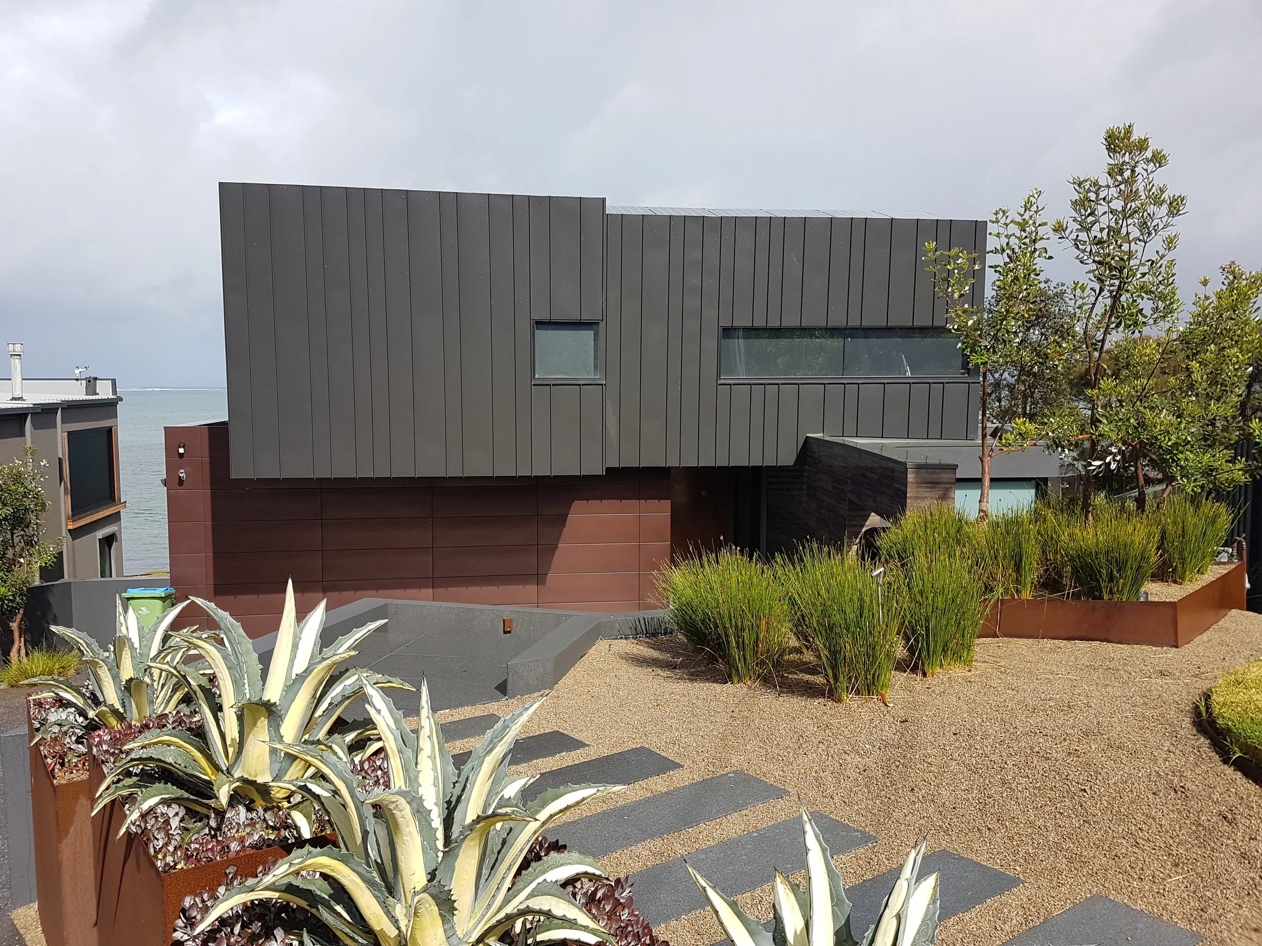Modern gray building with large windows, surrounded by desert plants and trees, with water and cloudy sky in the background.