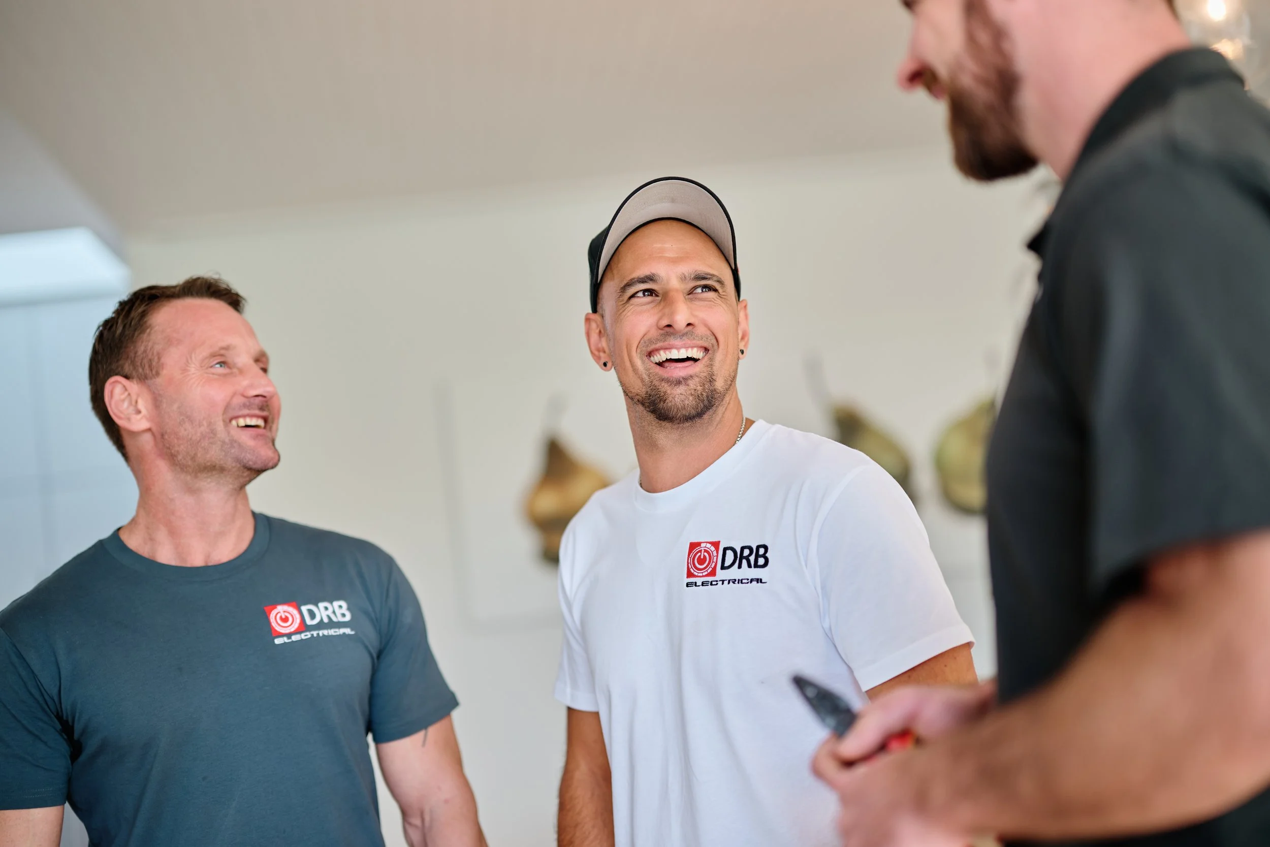 Three men smiling and talking to each other indoors. Two of them wear t-shirts with the logo of DRB Electrical; the man in the middle wears a cap. There is a blurred person in the foreground using a phone.