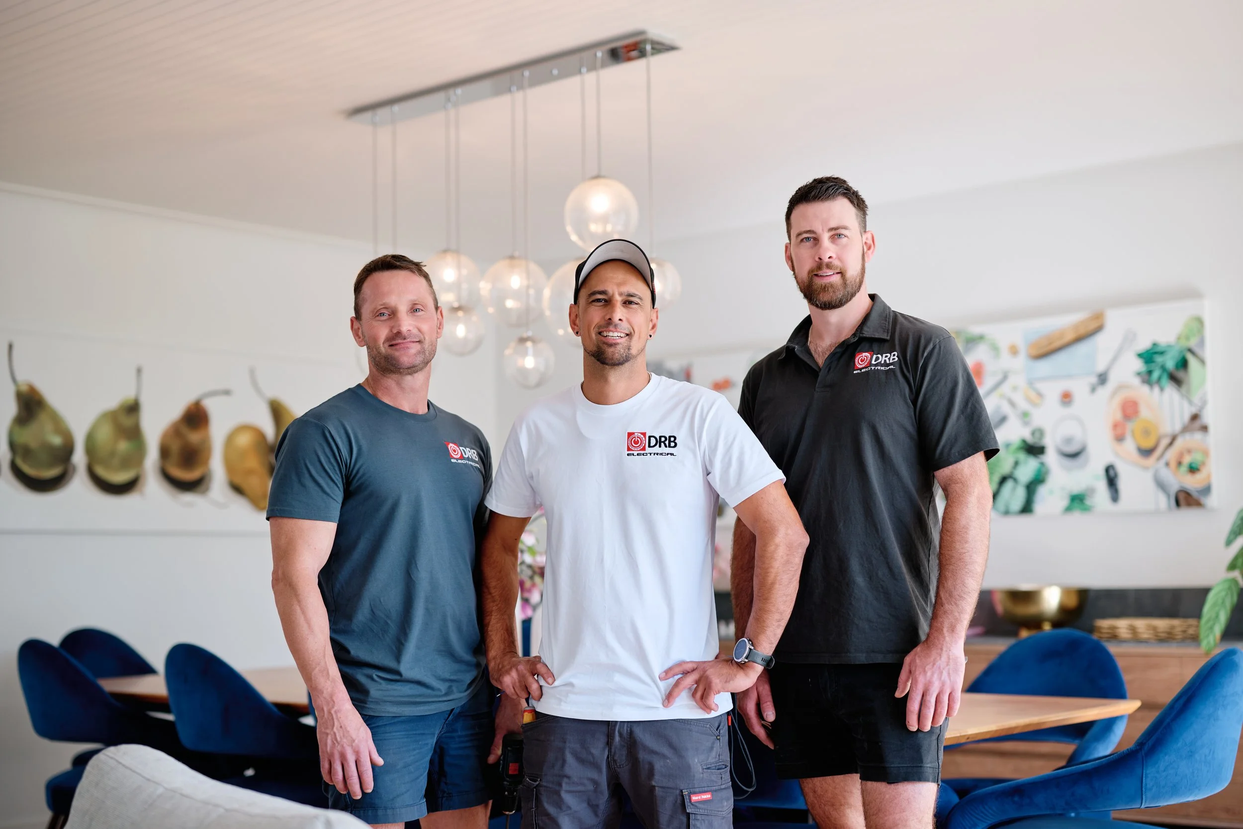 Three men standing together in a modern dining room, smiling, with a wall of hanging pears and kitchen utensils behind them.
