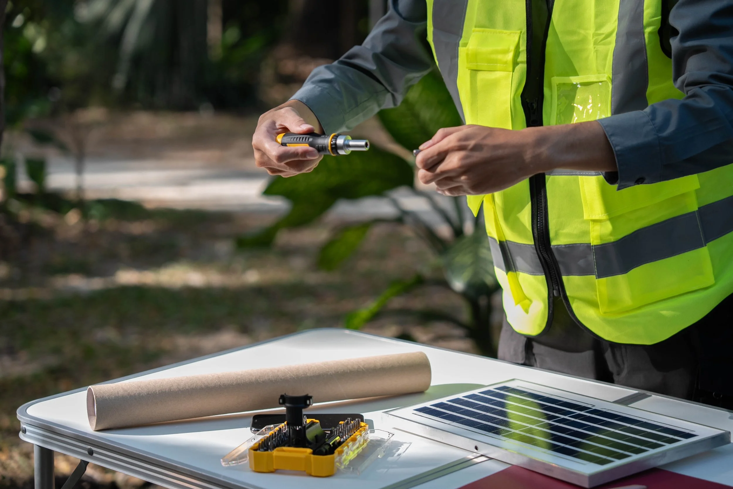 Close-up of a person wearing a yellow safety vest working with a small tool on a solar panel and other tools on a table outdoors.