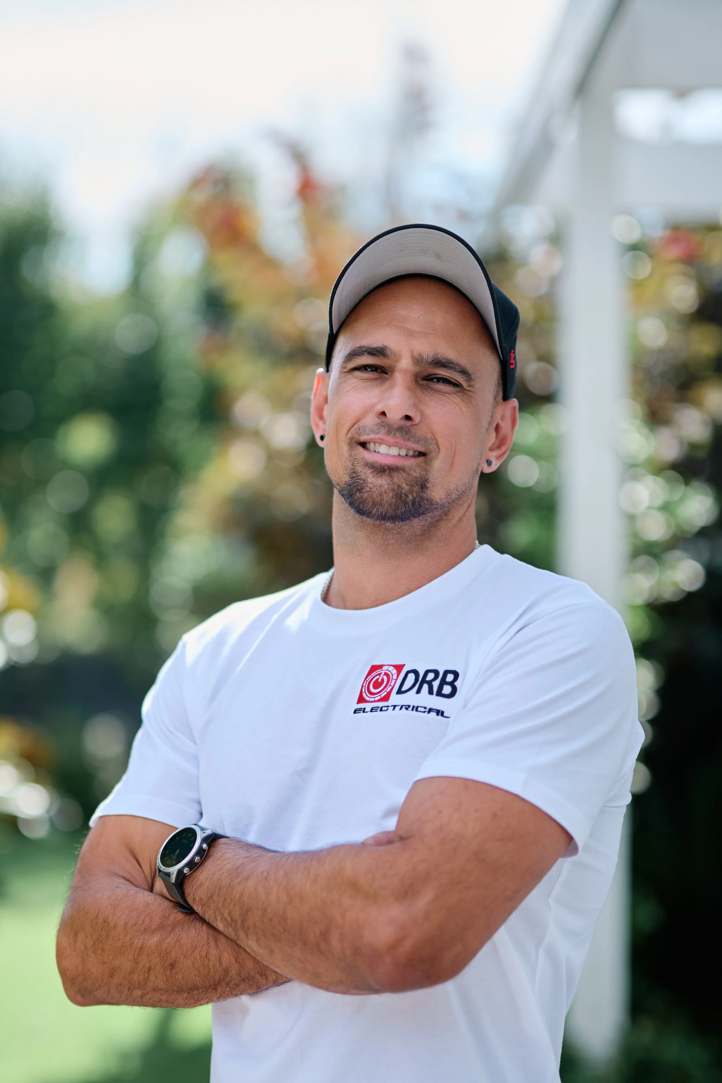 A man with a beard and earrings, wearing a white t-shirt with a logo that reads "DRB Electrical," a sports watch, a black cap, and standing outdoors with his arms crossed. The background is out of focus with trees and a structure.