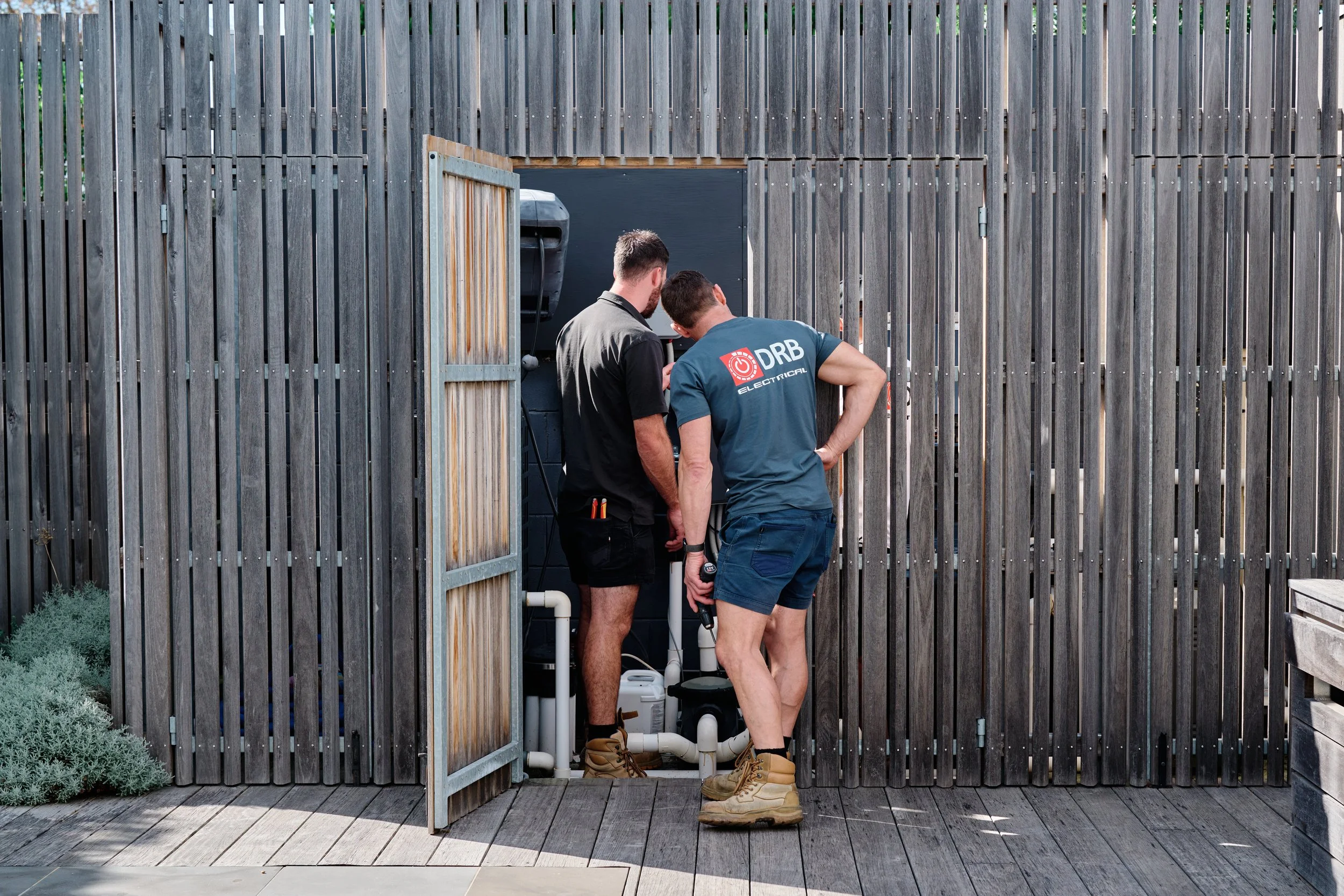 Two men working on an outdoor electrical or plumbing system behind a wooden fence and gate, wearing work boots and shirts, one with a Dr. Electrical logo.