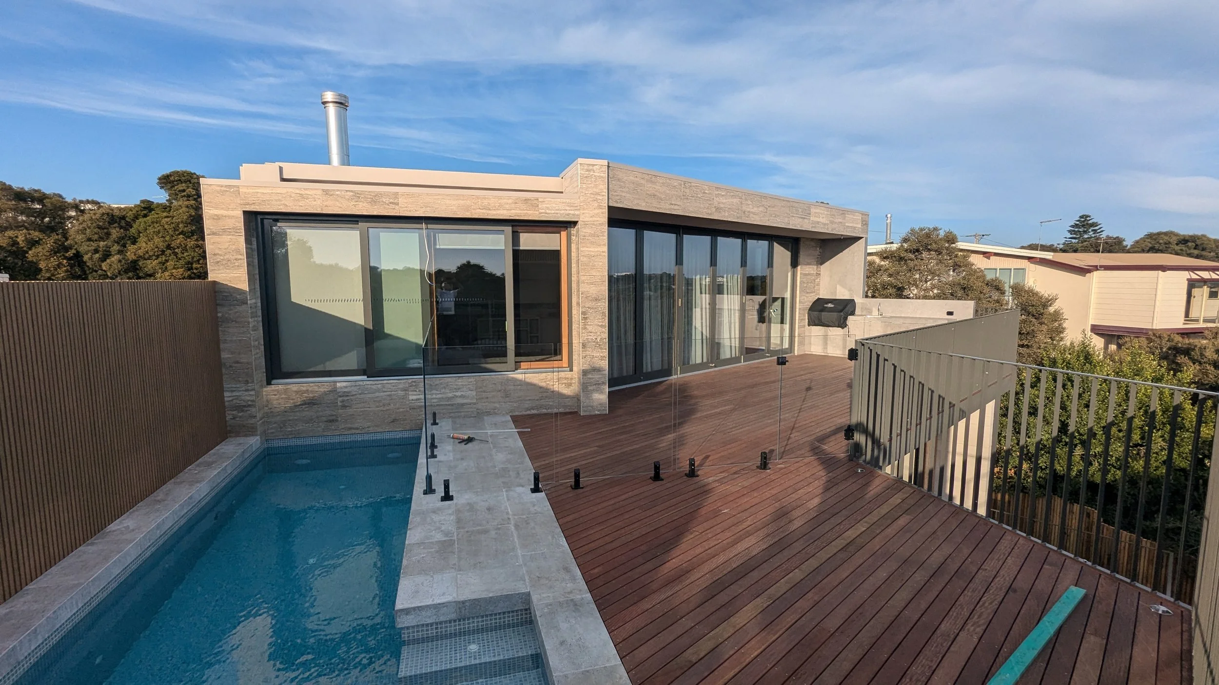 Modern house with large glass sliding doors, a small swimming pool, and a wooden deck, surrounded by metal and wooden fencing.