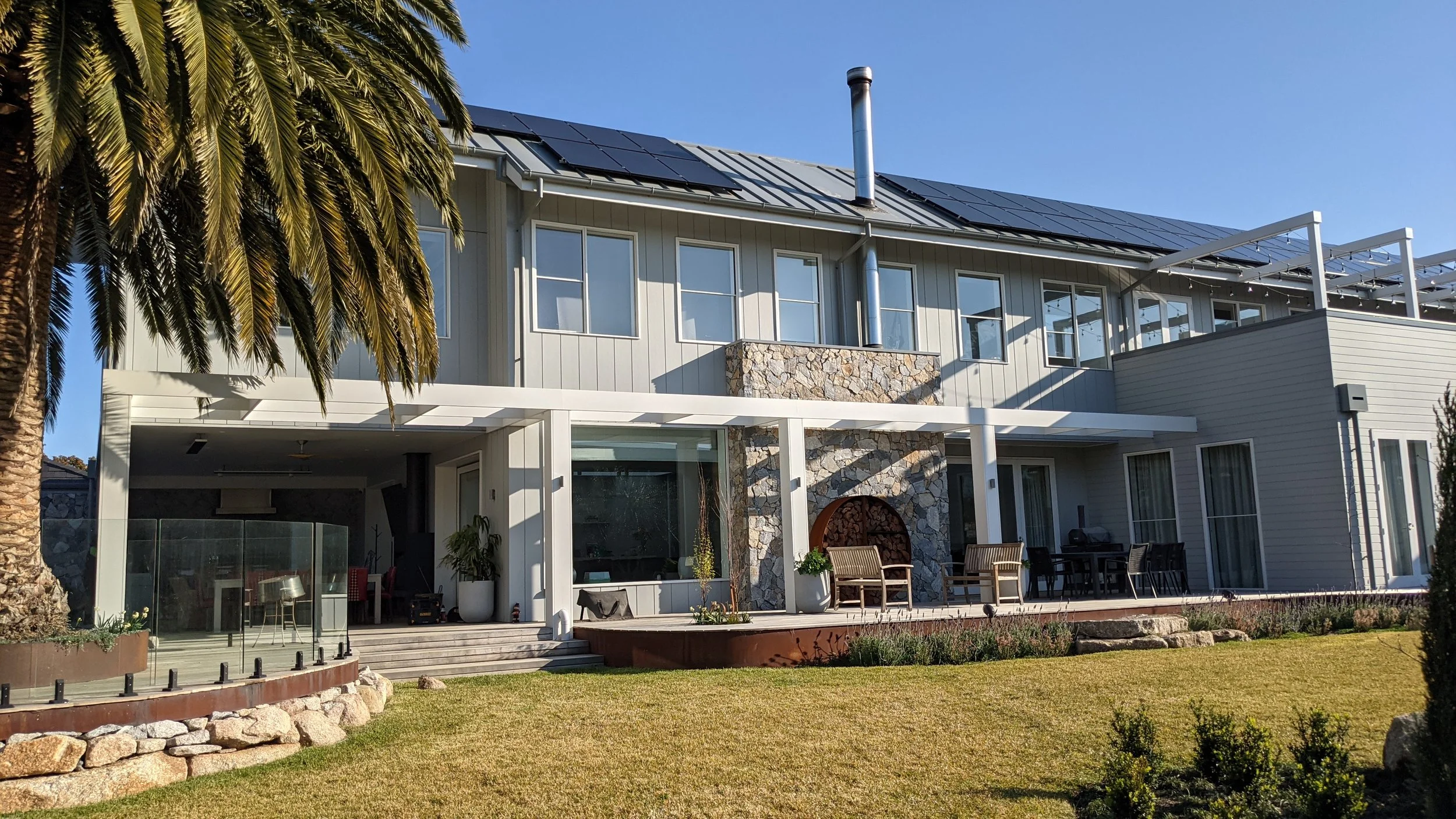 A modern two-story house with large windows, a stone accent wall, and solar panels on the roof, with a palm tree and a lawn in the foreground under a clear blue sky.