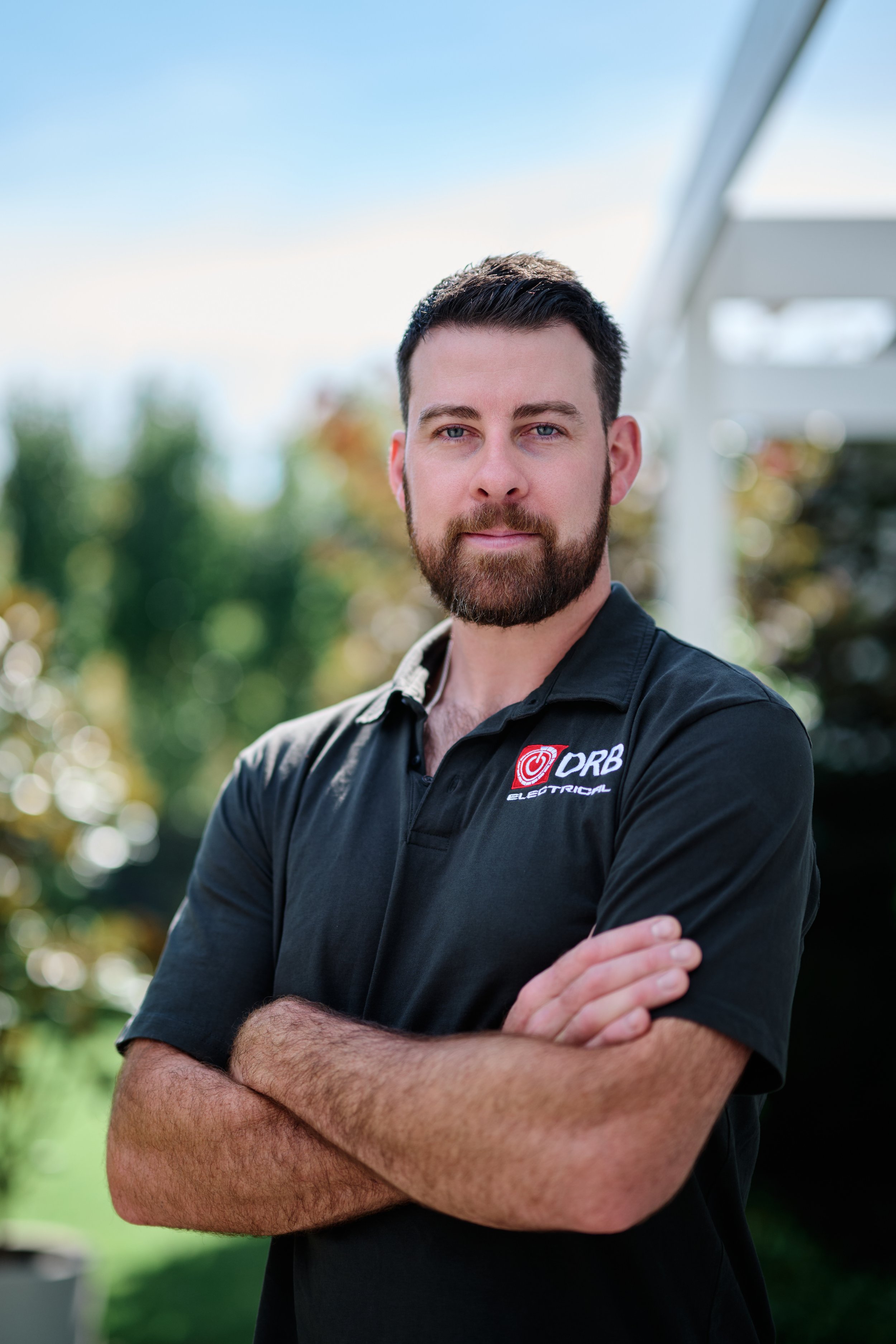 A man with dark hair and a beard, wearing a black collared shirt with a logo that reads "DRB Electrical," stands outdoors with his arms crossed, blurred trees in the background, under a blue sky.