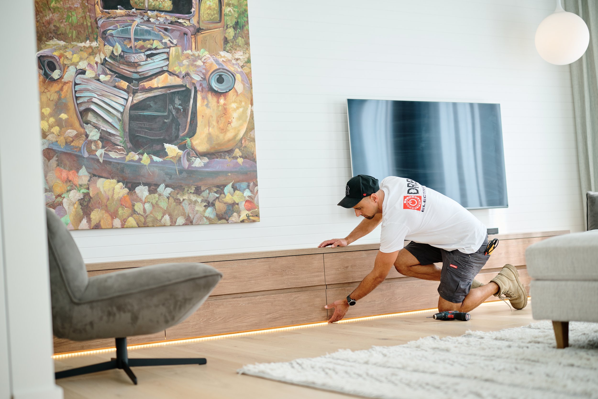 A man is kneeling on the floor installing or inspecting a wooden baseboard with integrated lighting in a modern living room. Behind him, a large colorful painting of an abandoned vintage car is hung on a white wall, which features horizontal paneling. There is a flat-screen TV mounted on the wall and a white round ceiling light fixture visible in the room.