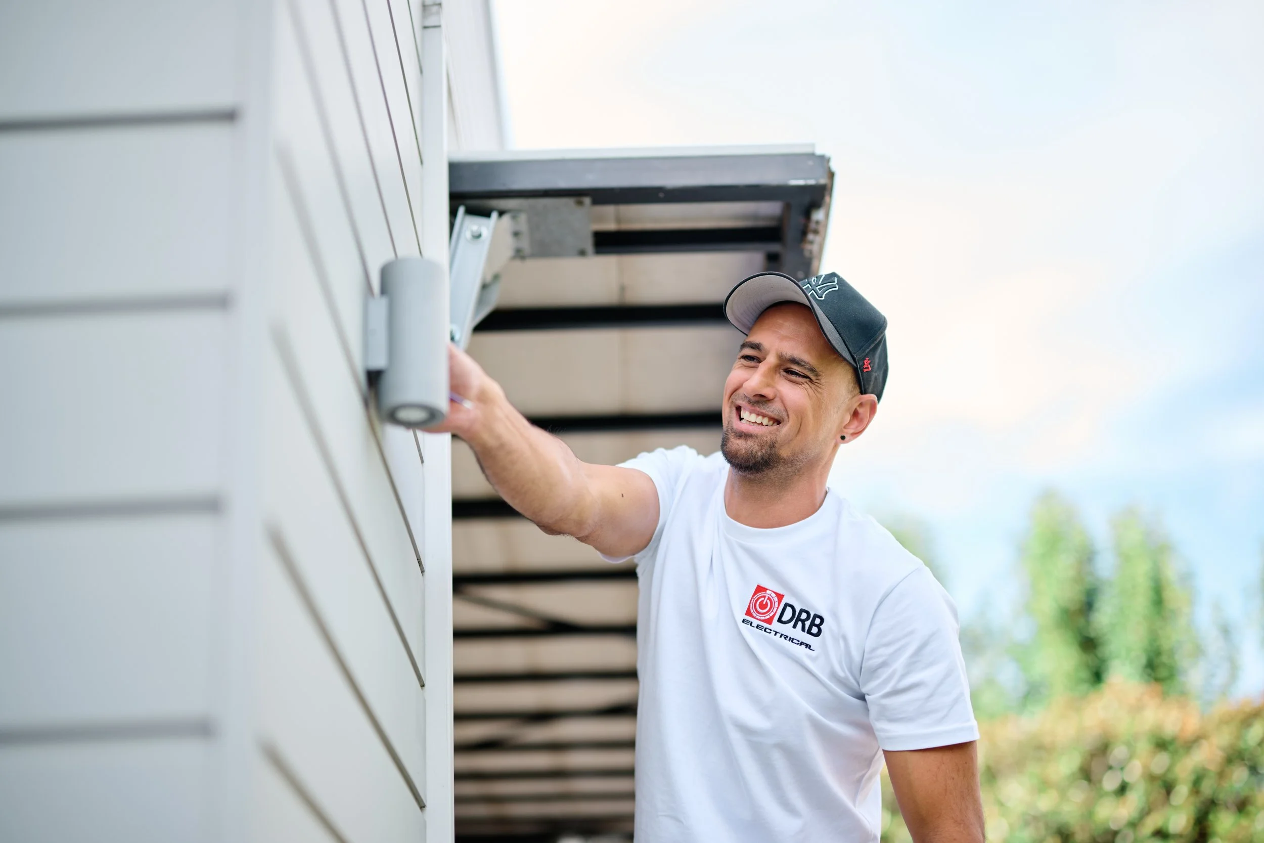 A man in a white t-shirt and a black baseball cap is installing or repairing an outdoor electrical or communication device on the side of a building with horizontal siding. He is smiling and appears to be working on the device with his right hand.