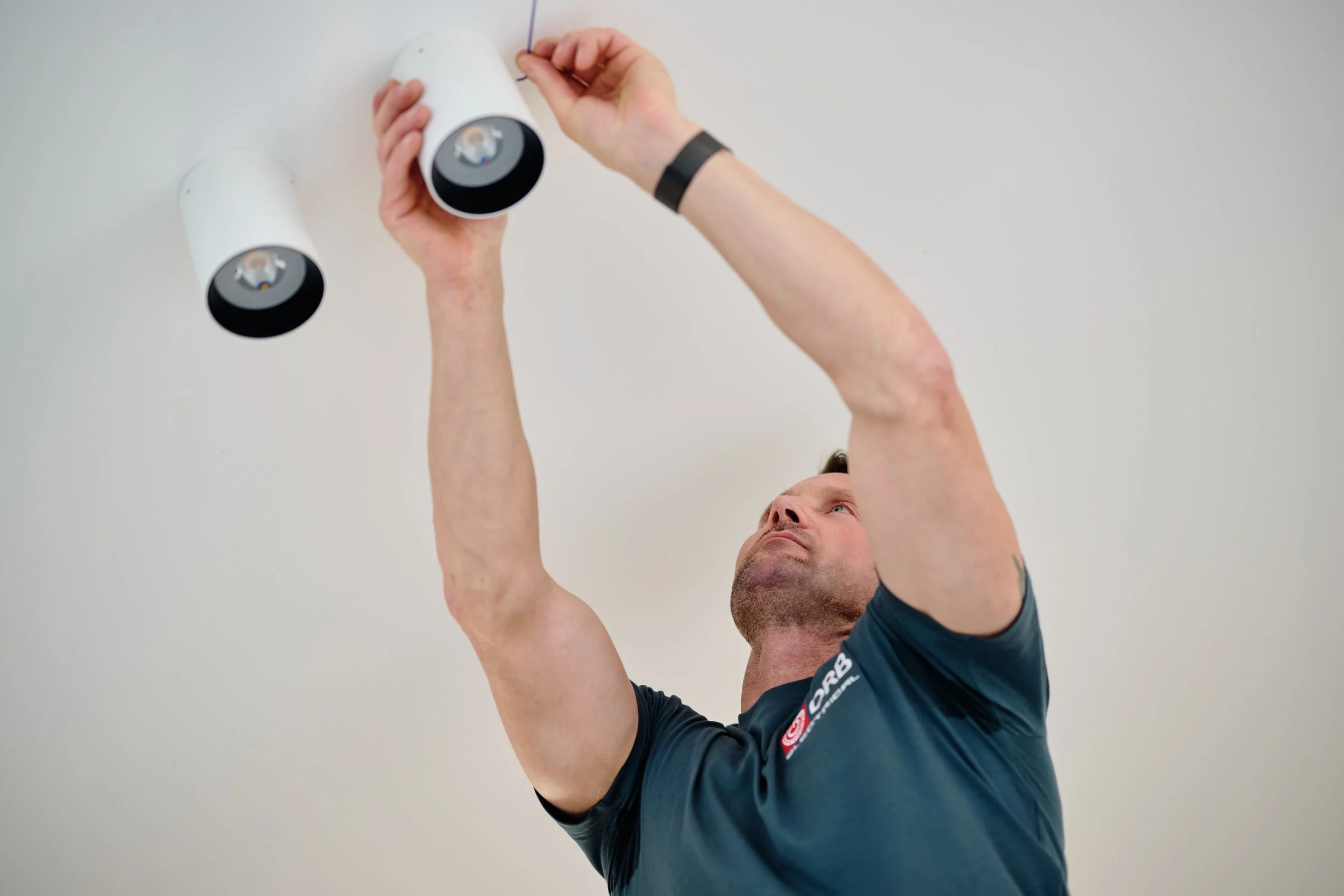 A man with short hair and beard, wearing a dark T-shirt with logos, is installing a smoke detector on a white ceiling. He is holding the detector with one hand and using the other hand to screw it in.