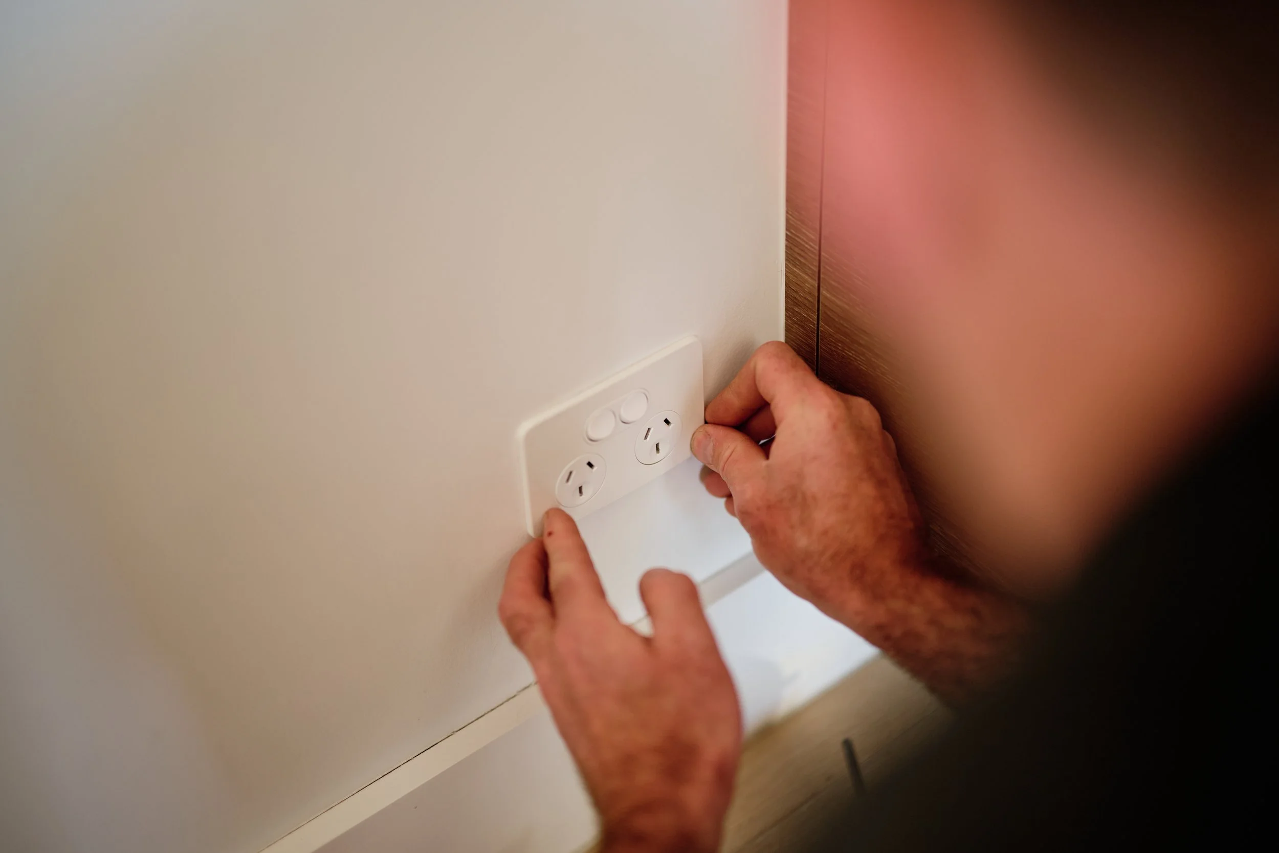 Close-up of hands installing an electrical outlet on a white wall next to a wooden surface.