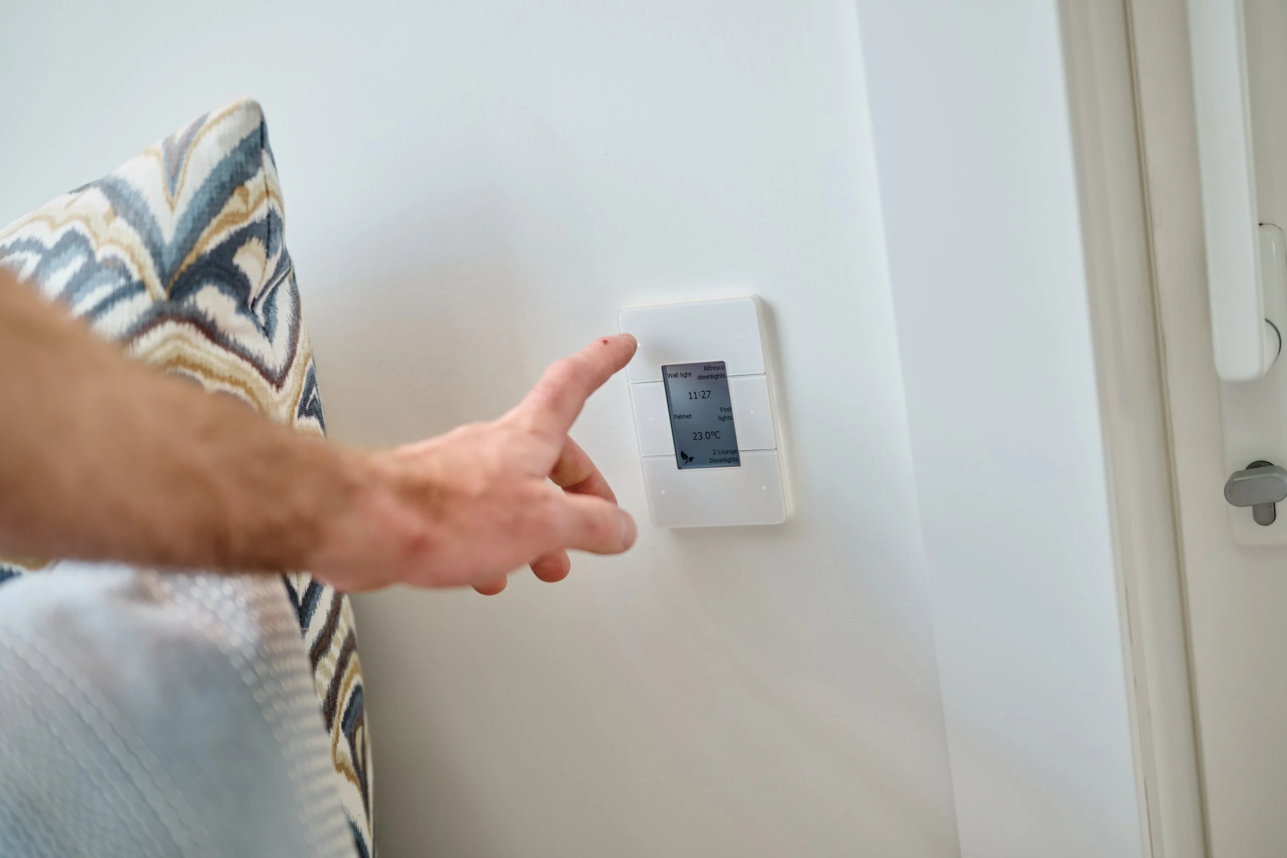Person tapping a digital wall control panel on a white wall, with a patterned curtain partially visible on the left.