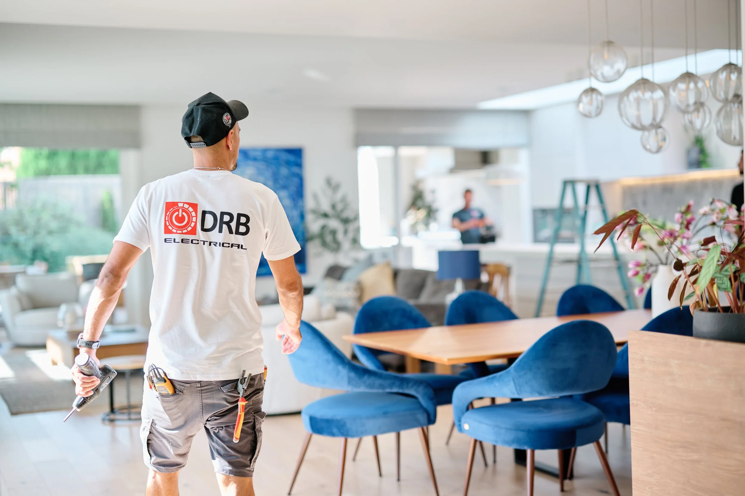 A man with a black cap and a white t-shirt labeled 'DRB Electrical' is working in a modern, well-lit living or dining area. He is holding a power drill in his right hand and has tools attached to his waistband. In the background, another person is visible, along with contemporary furniture and decorative light fixtures hanging from the ceiling.