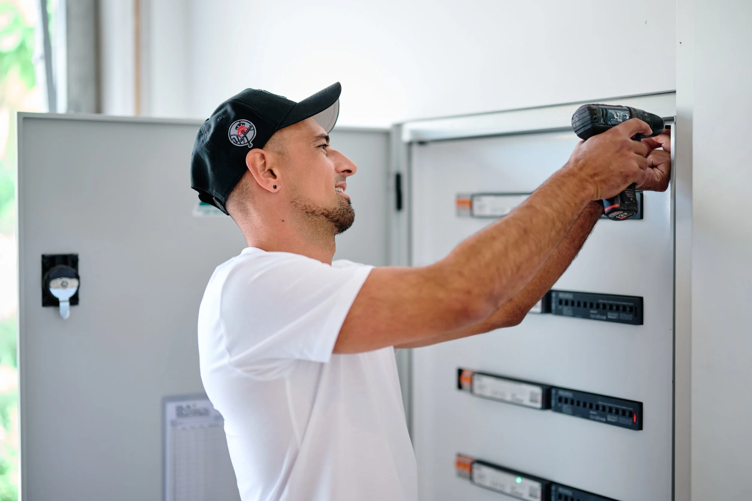 A man wearing a black baseball cap and white t-shirt is working with electrical tools on an electrical panel or circuit breaker box inside a building.