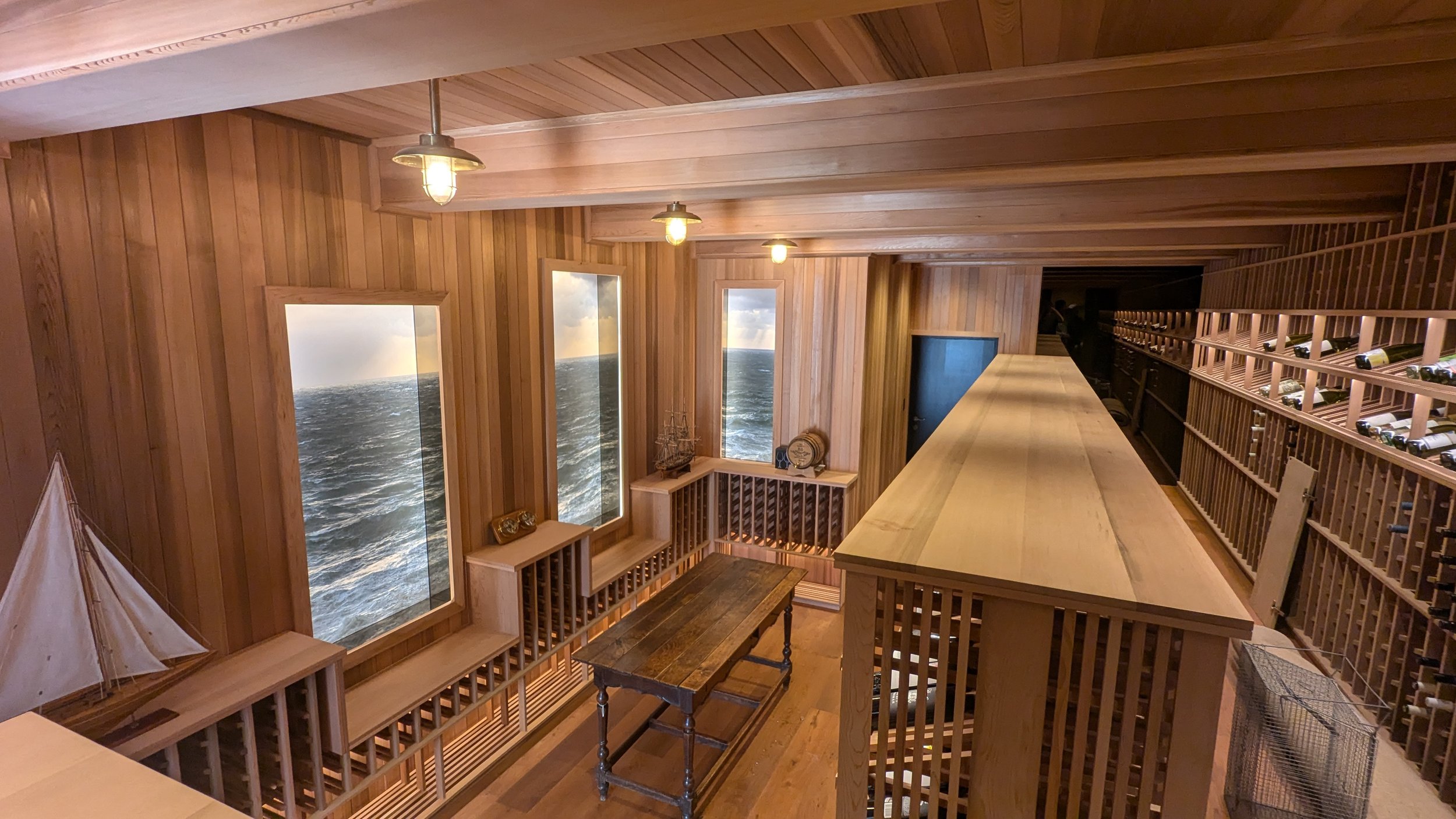 A cozy wooden-paneled room with large ocean-view windows showing waves and cloudy sky. There is a long countertop, wine racks filled with bottles, and nautical decorations including a model ship and a small sailboat.