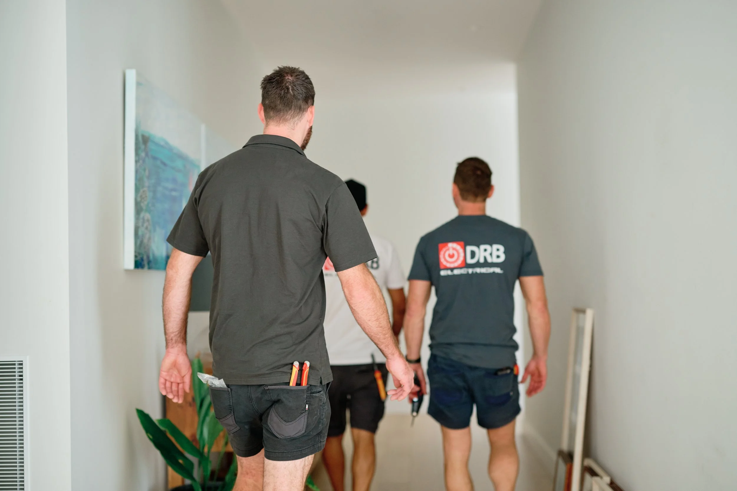 Three men walking down a corridor, with the man in the foreground wearing a gray shirt and shorts, carrying tools in his pocket, and the other two in blue shirts with logos and text on the back.