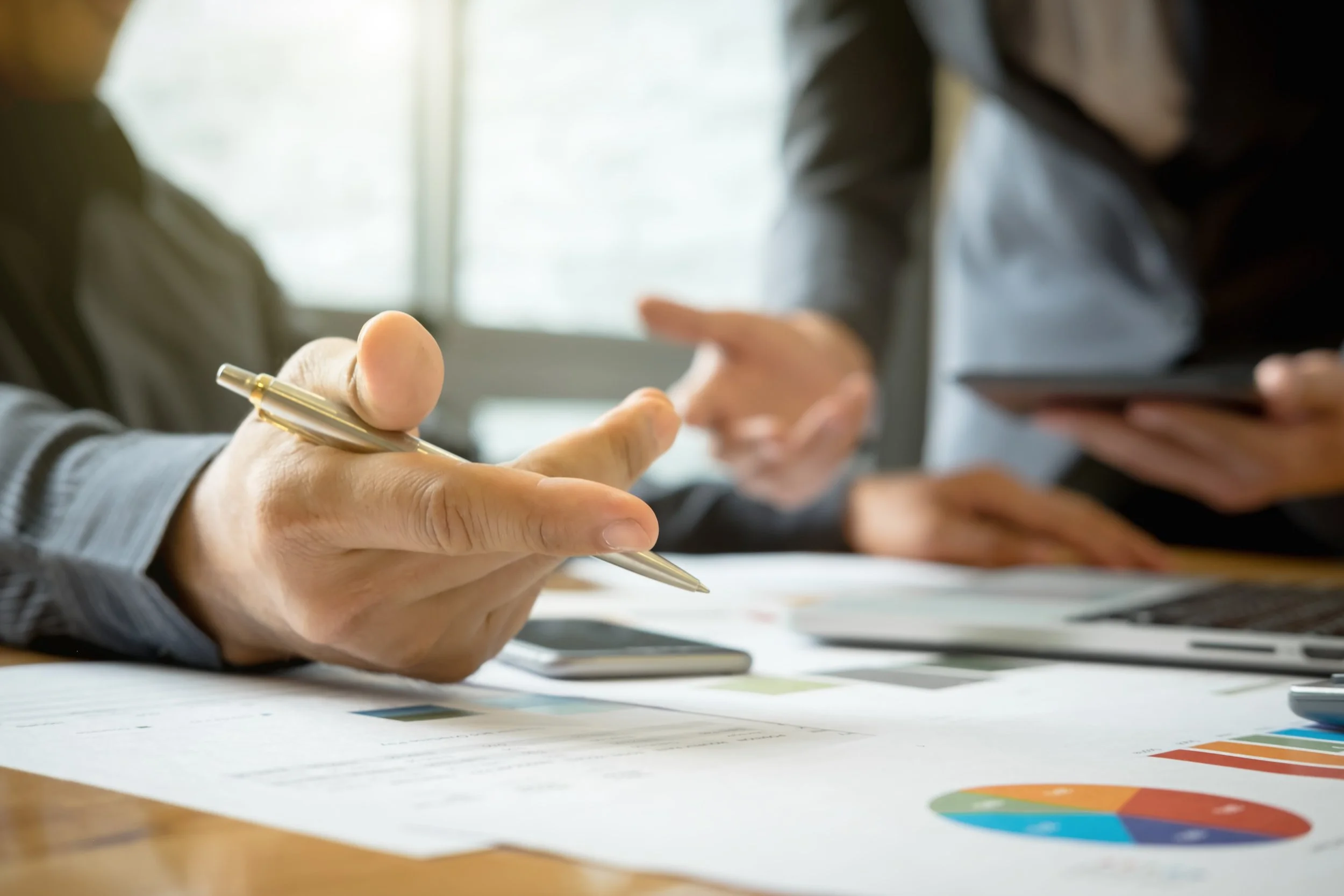 Close-up of a person's hand holding a gold-colored pen, writing on papers with charts and graphs, with two people in business attire engaged in a discussion in the blurred background.