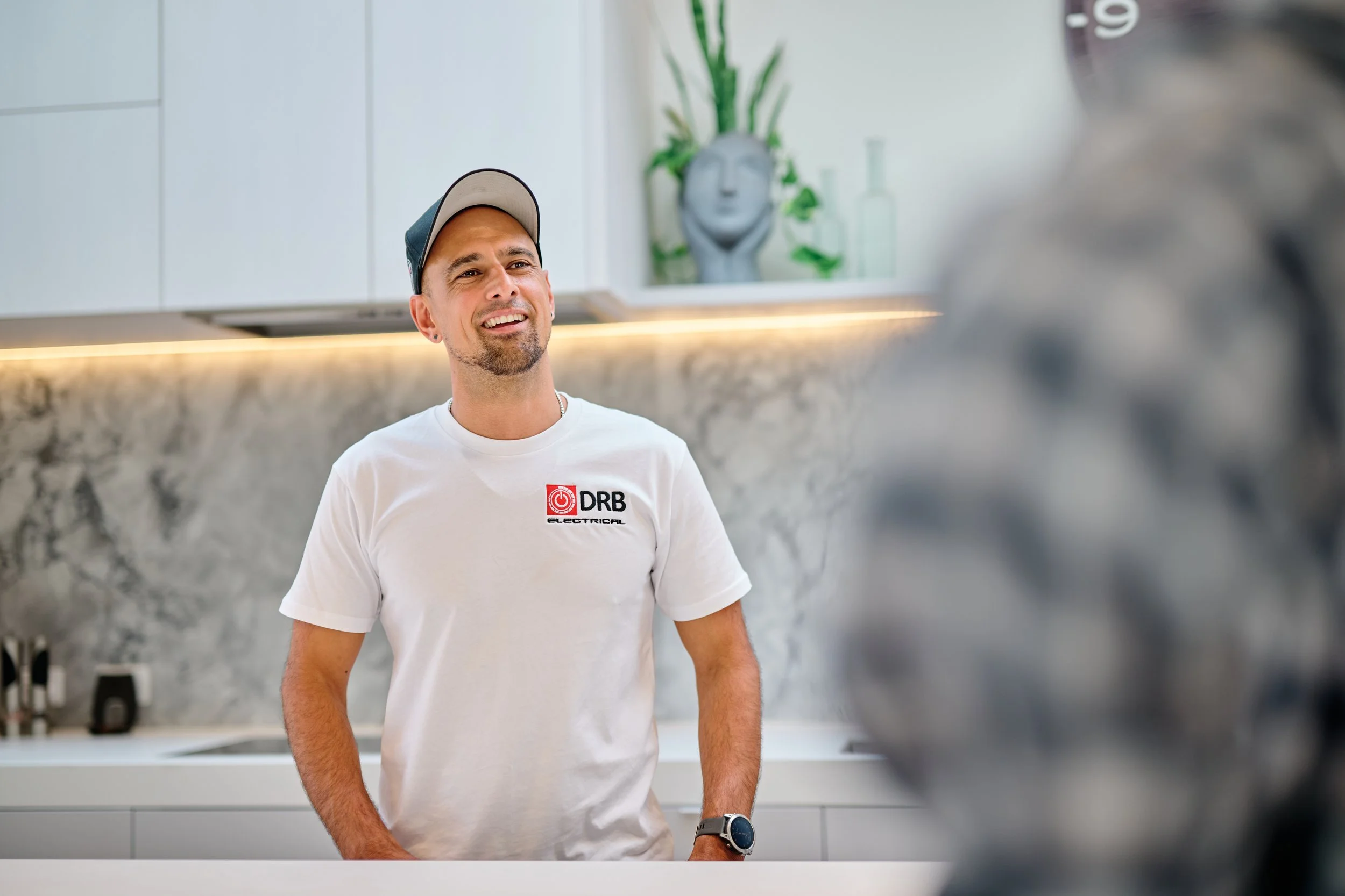 Man wearing a white t-shirt with a logo and a gray cap, standing in a modern kitchen, smiling and looking at someone or something off-camera.
