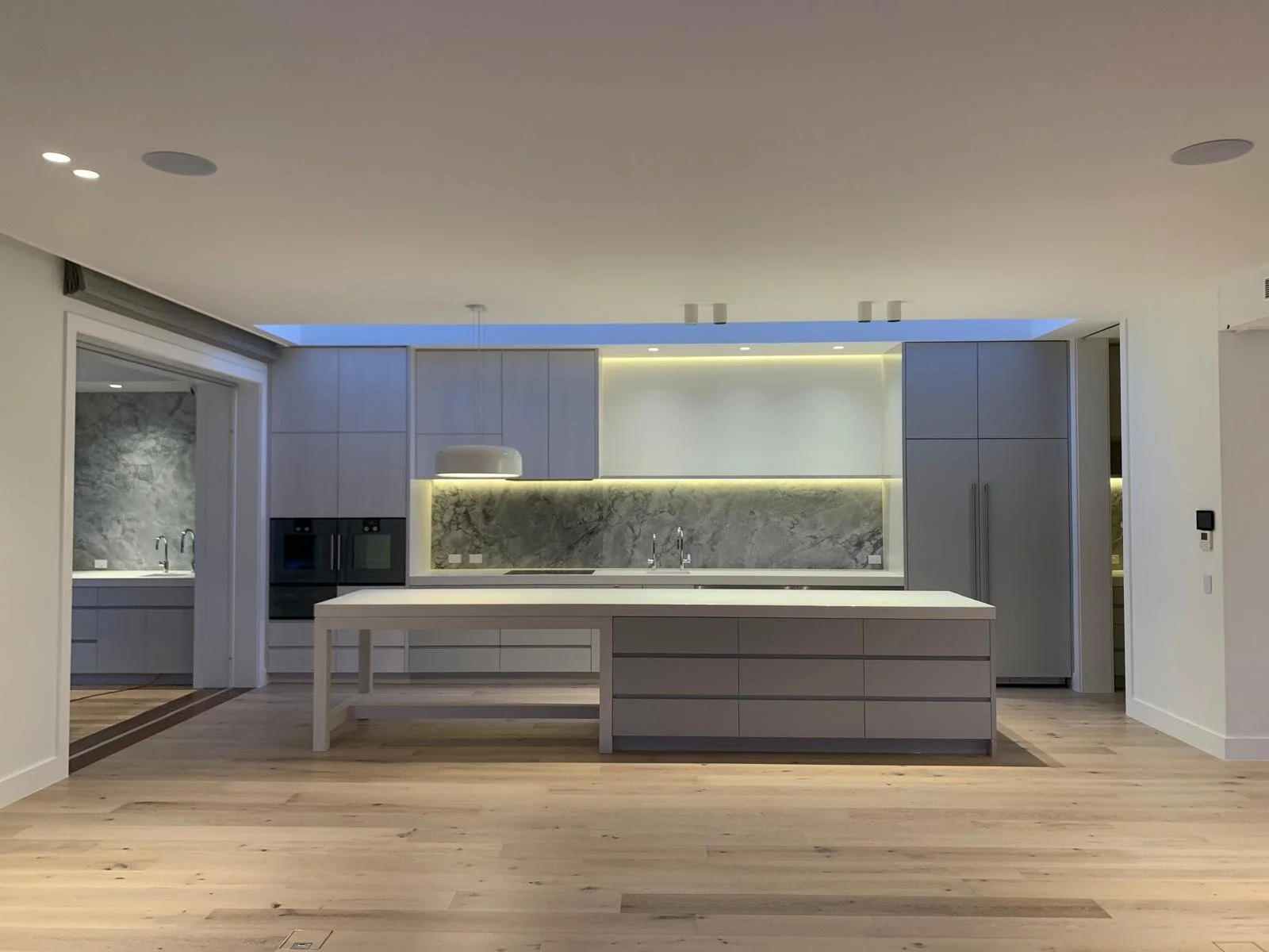 Modern kitchen with white cabinetry, marble backsplash, and a large island in the center, illuminated by warm and cool lighting.