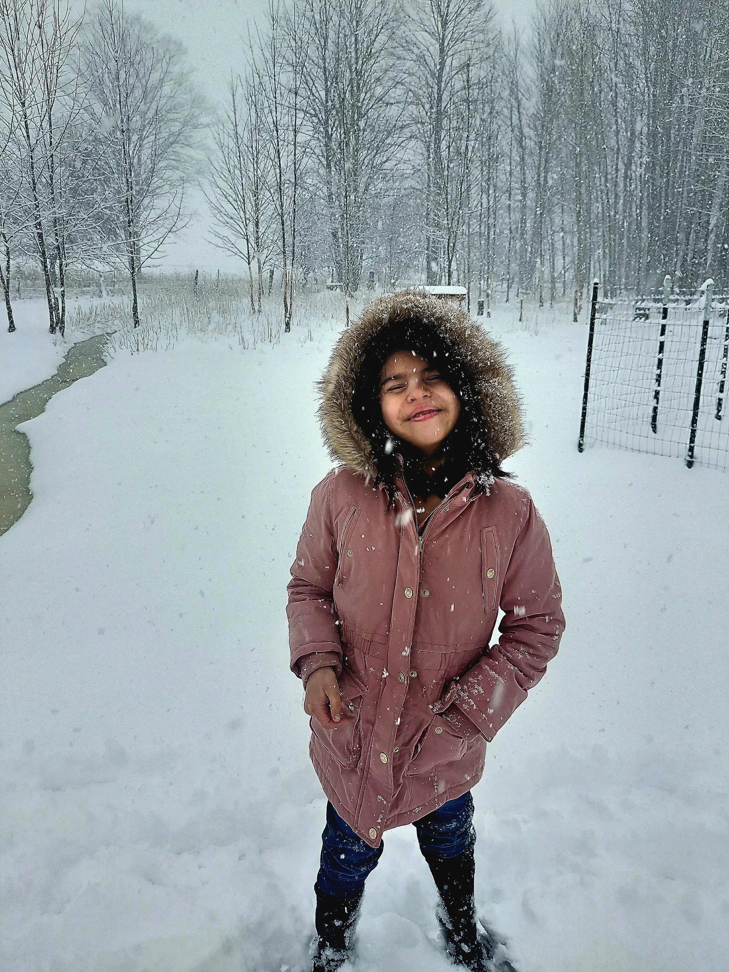 A smiling girl wearing a pink winter coat with a fur-lined hood standing in the snow during a snowy day surrounded by trees and a fence.