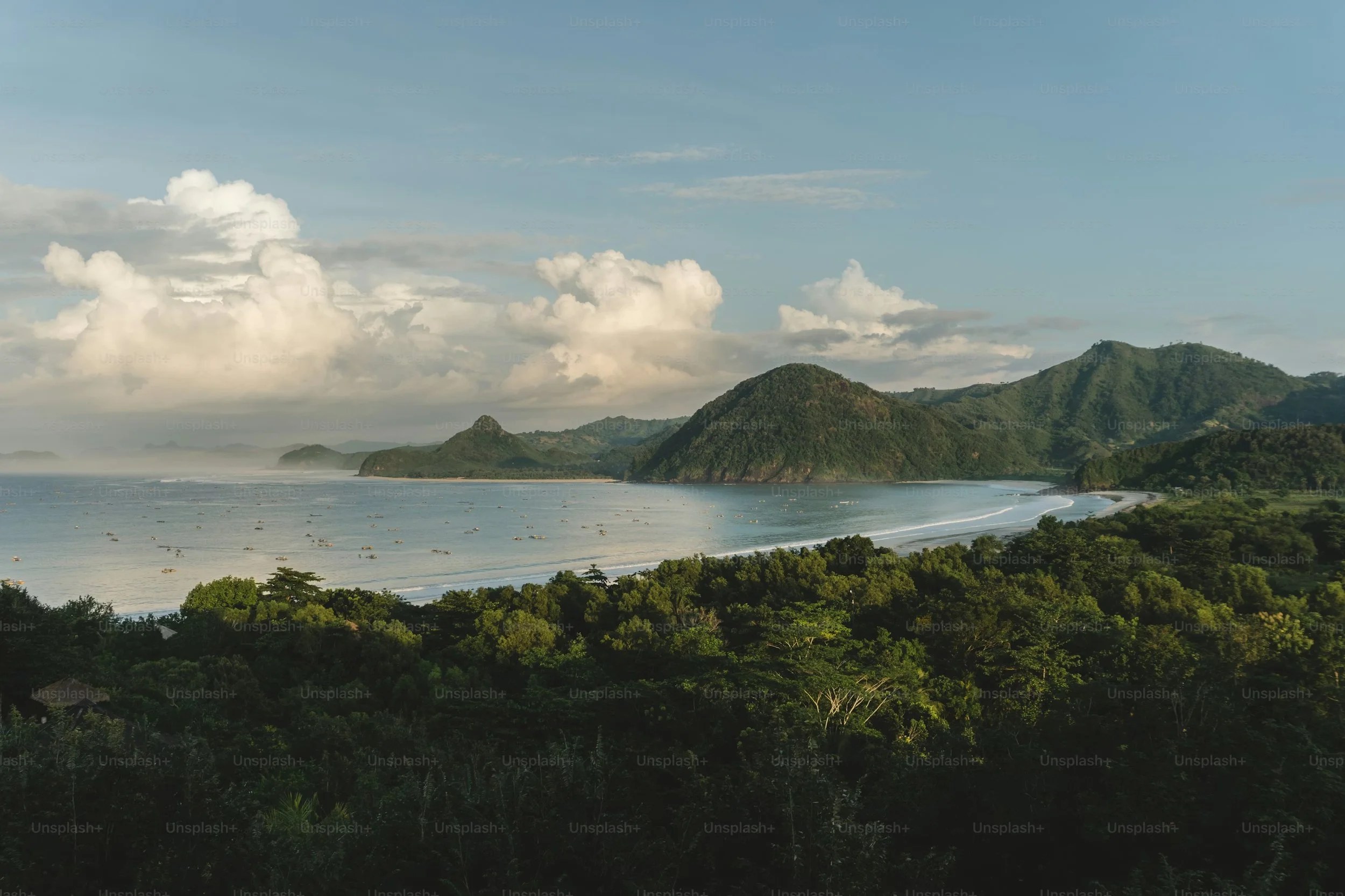 Paysage côtier avec des montagnes verdoyantes, une plage de sable clair et des nuages blancs dans le ciel.