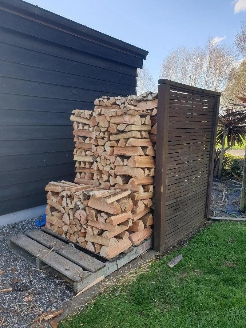 Stack of firewood logs outside a black shed with a wooden privacy fence on the right side, on a small pallet, with grass and some plants nearby.