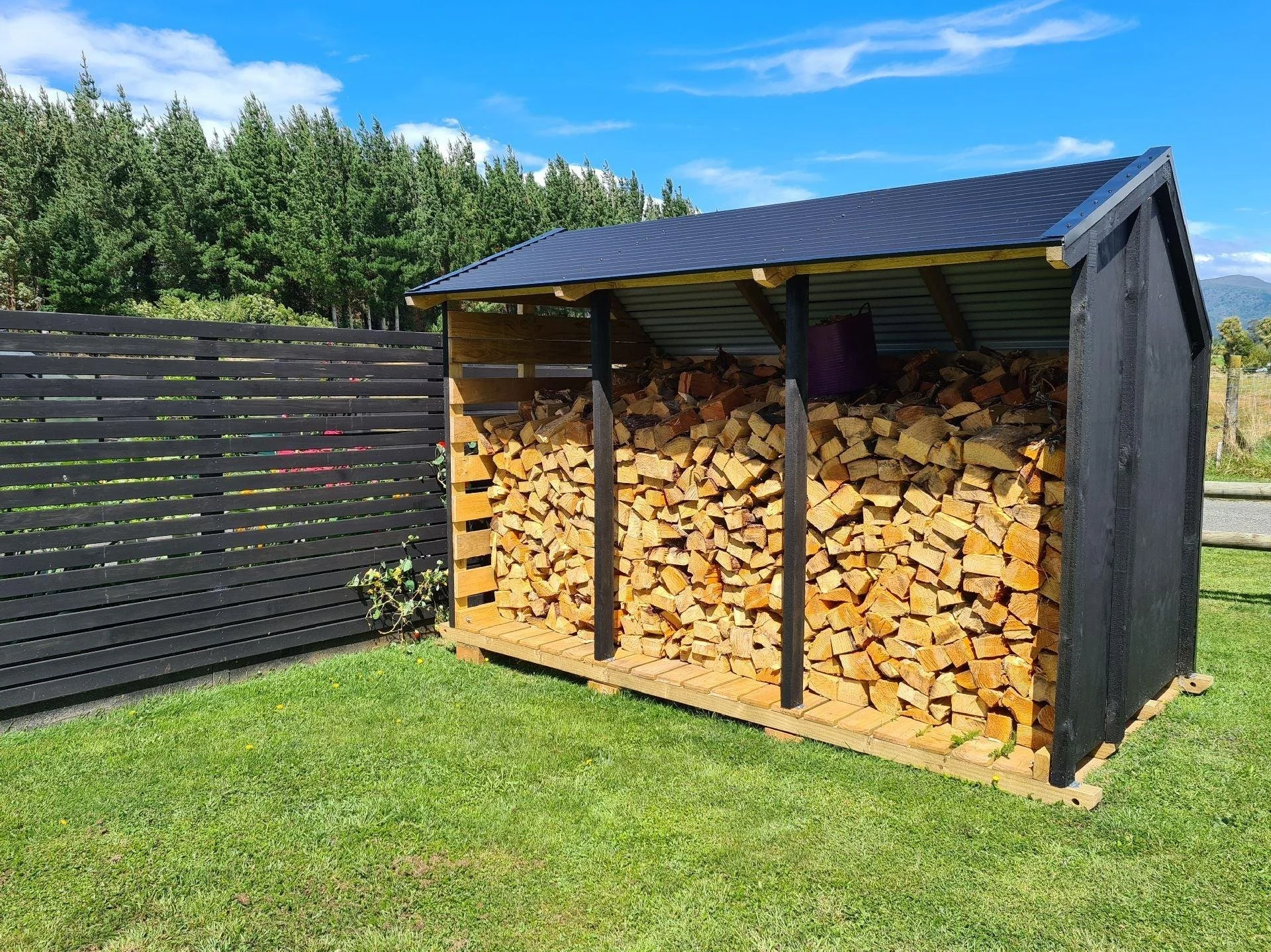 Woodpile stored in a black wooden shed with a metal roof, outdoors on green grass with trees and a blue sky in the background.