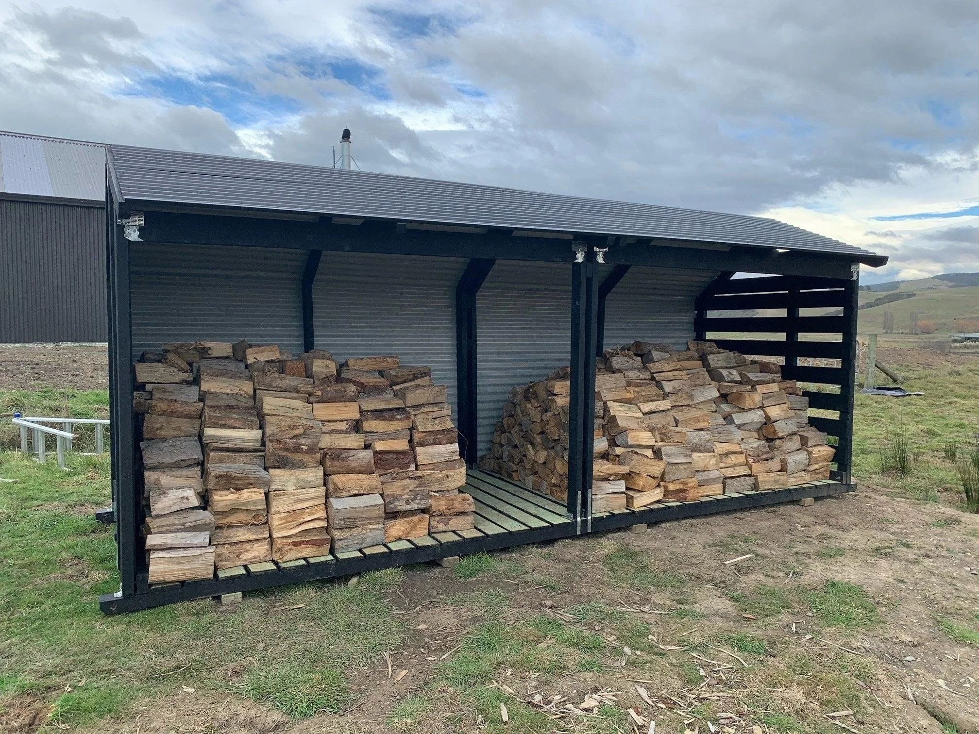 A metal wood storage shed with an open side, filled with stacked firewood, situated in a rural outdoor landscape under a partly cloudy sky.