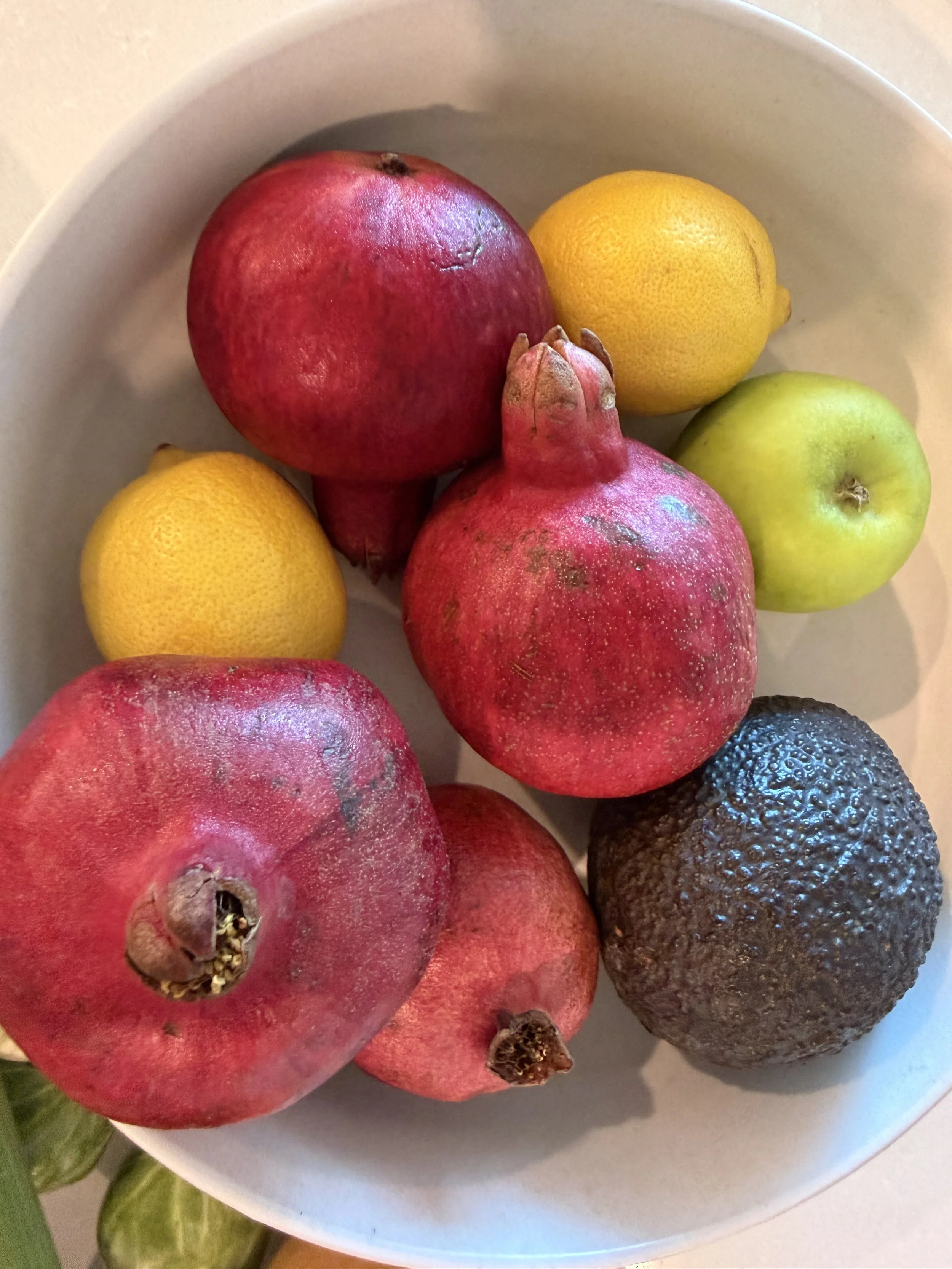 A white bowl containing red pomegranates, green apples, yellow lemons, and a dark purple avocado.