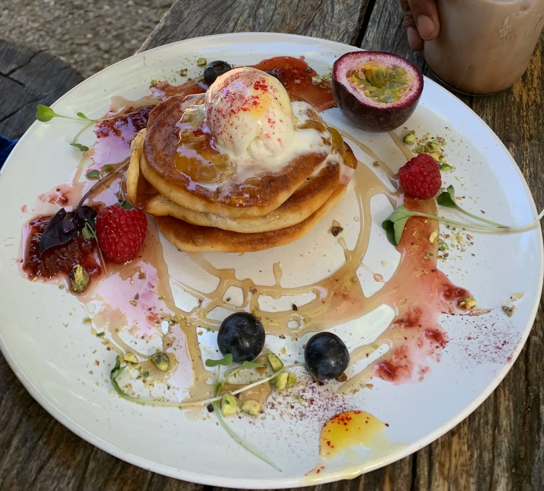 A partially eaten pancake topped with a scoop of ice cream, served with assorted berries including raspberries and blueberries, and garnished with microgreens, syrup, and various sauces on a white plate on a rustic wooden table.