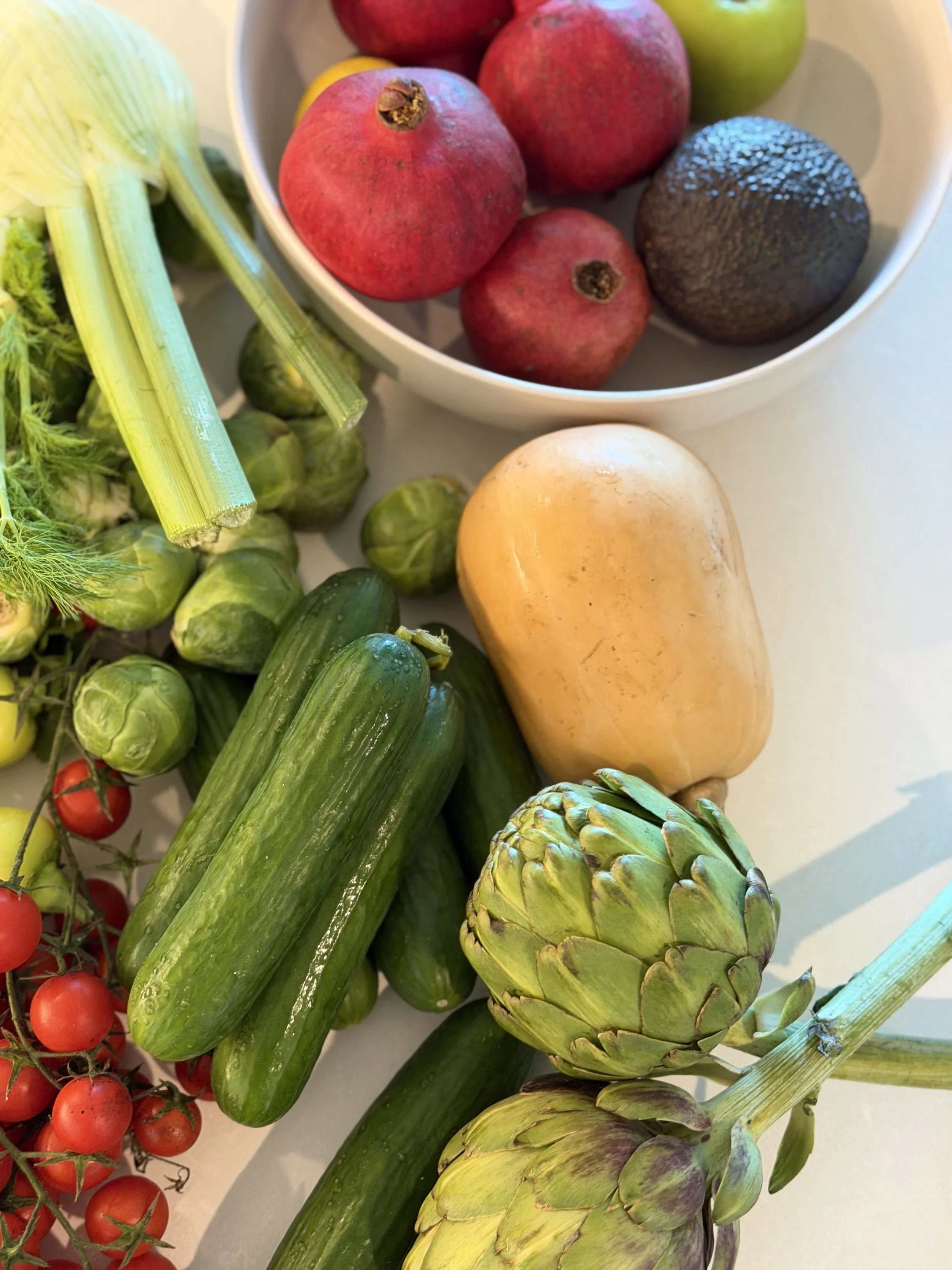 Assorted fresh vegetables and fruits, including cherry tomatoes, zucchinis, artichokes, a butternut squash, and pomegranates, arranged on a white surface.