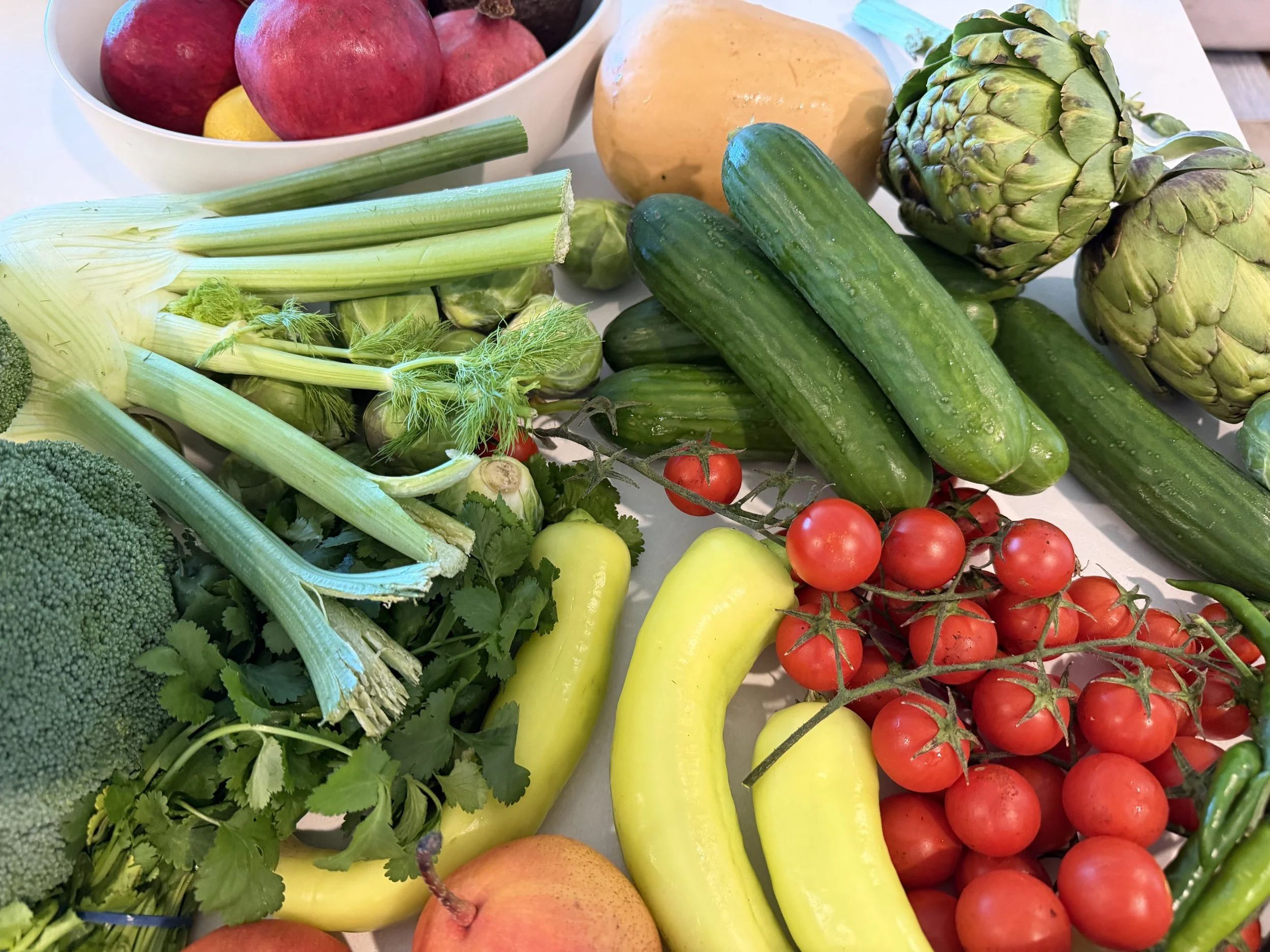 A colorful assortment of fresh fruits and vegetables including broccoli, celery, cherry tomatoes, green cucumbers, yellow zucchini, artichokes, an orange, apples, and a bowl of pomegranate seeds.