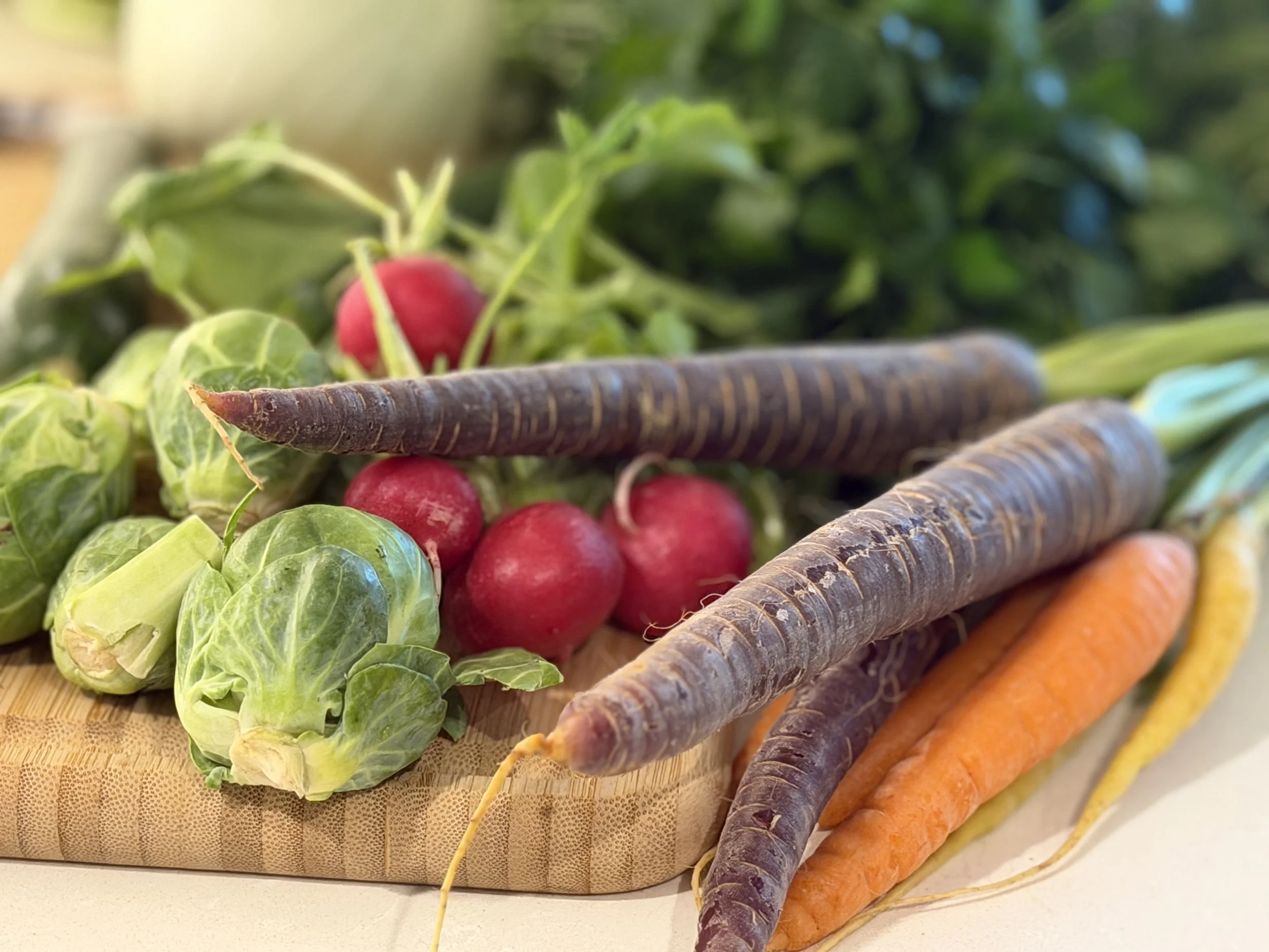 An assortment of fresh vegetables including Brussels sprouts, radishes, purple carrots, and orange carrots, arranged on a cutting board.