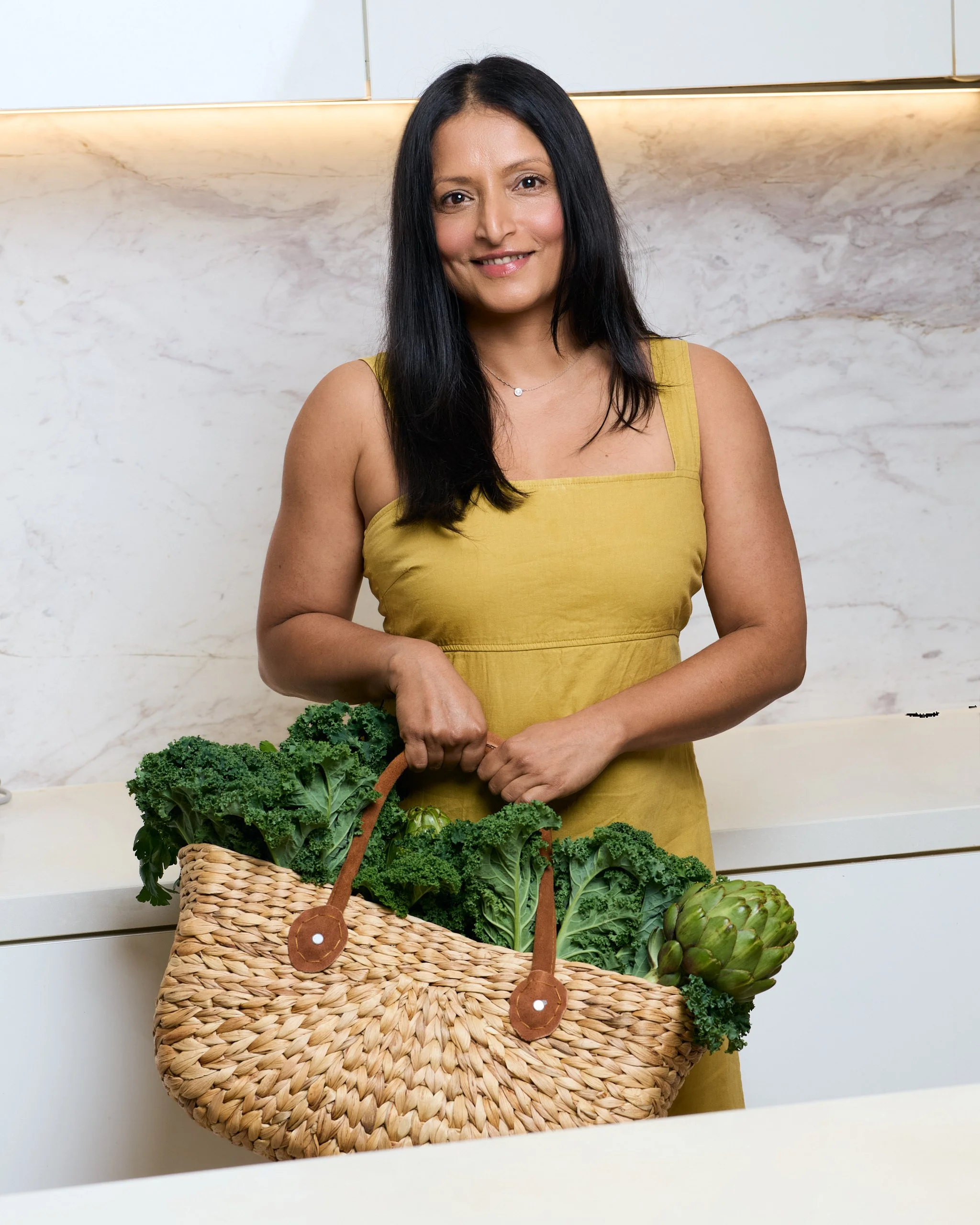 A woman with long black hair wearing a yellow dress standing behind a basket of green vegetables, including kale and artichokes, in a modern kitchen.