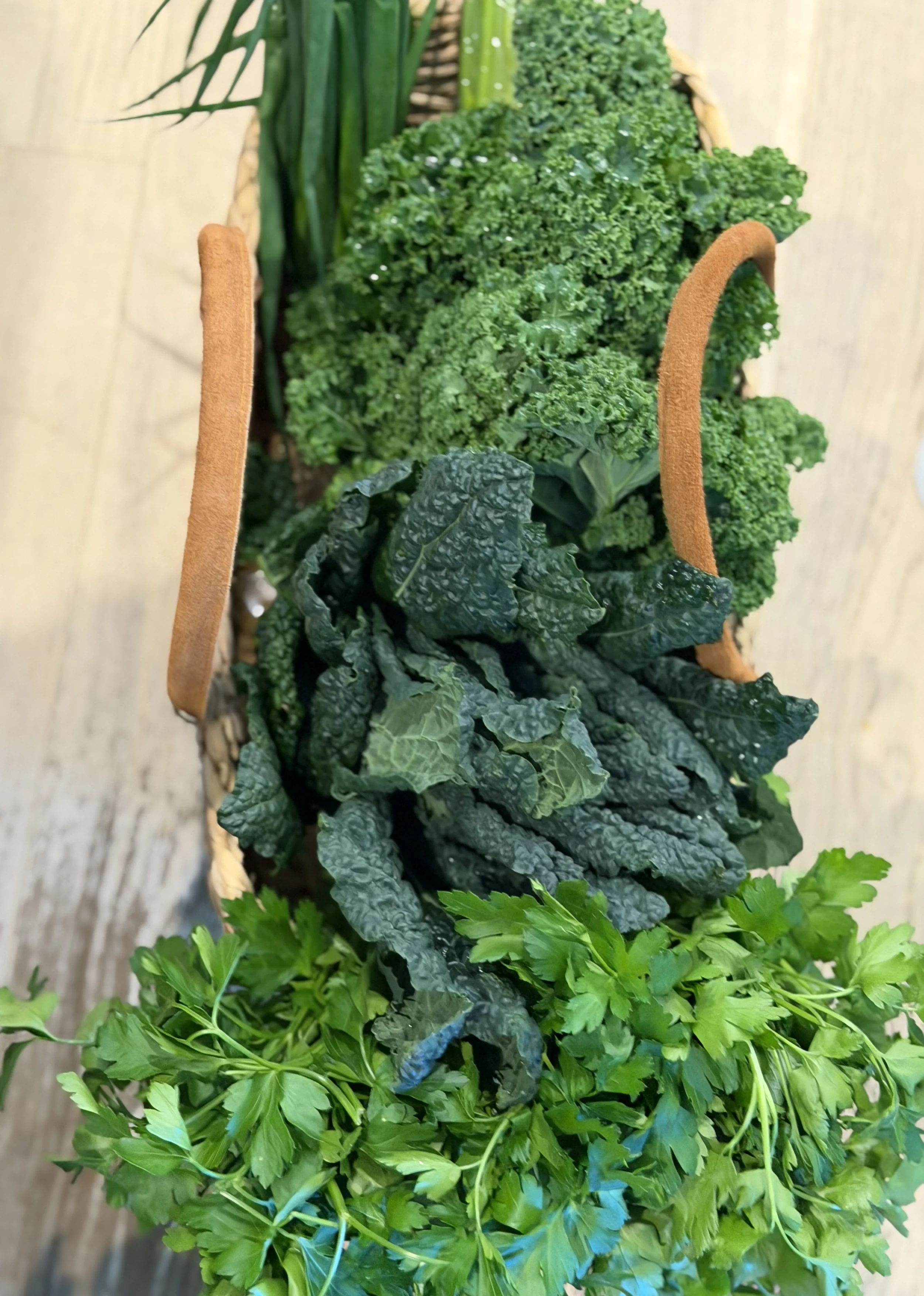 A basket with fresh green curly kale, flat-leaf parsley, and possibly chives or green onions on a wooden surface.