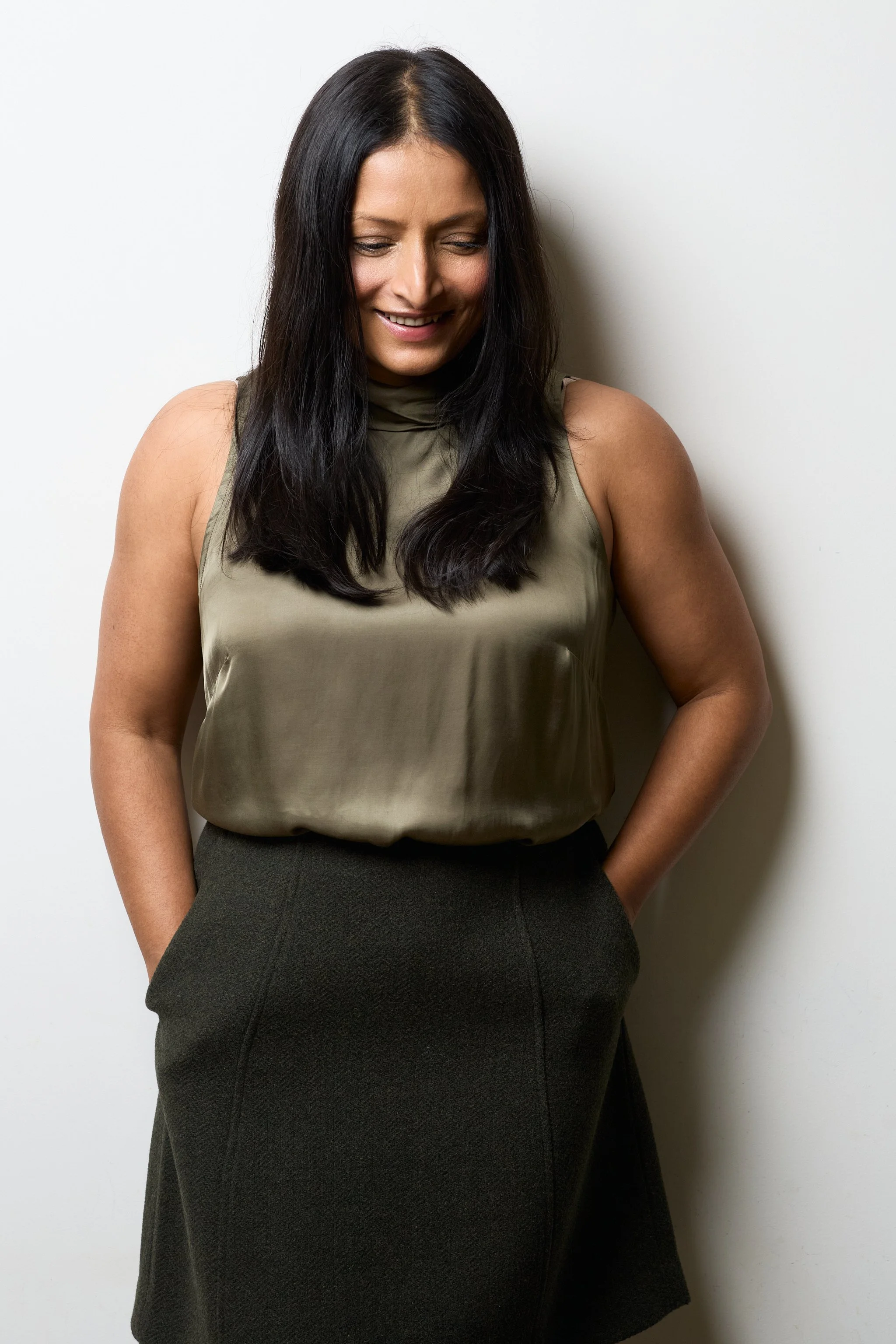 A woman with long black hair wearing a sleeveless beige top and a dark high-waisted skirt, standing against a plain white wall, looking down and smiling.