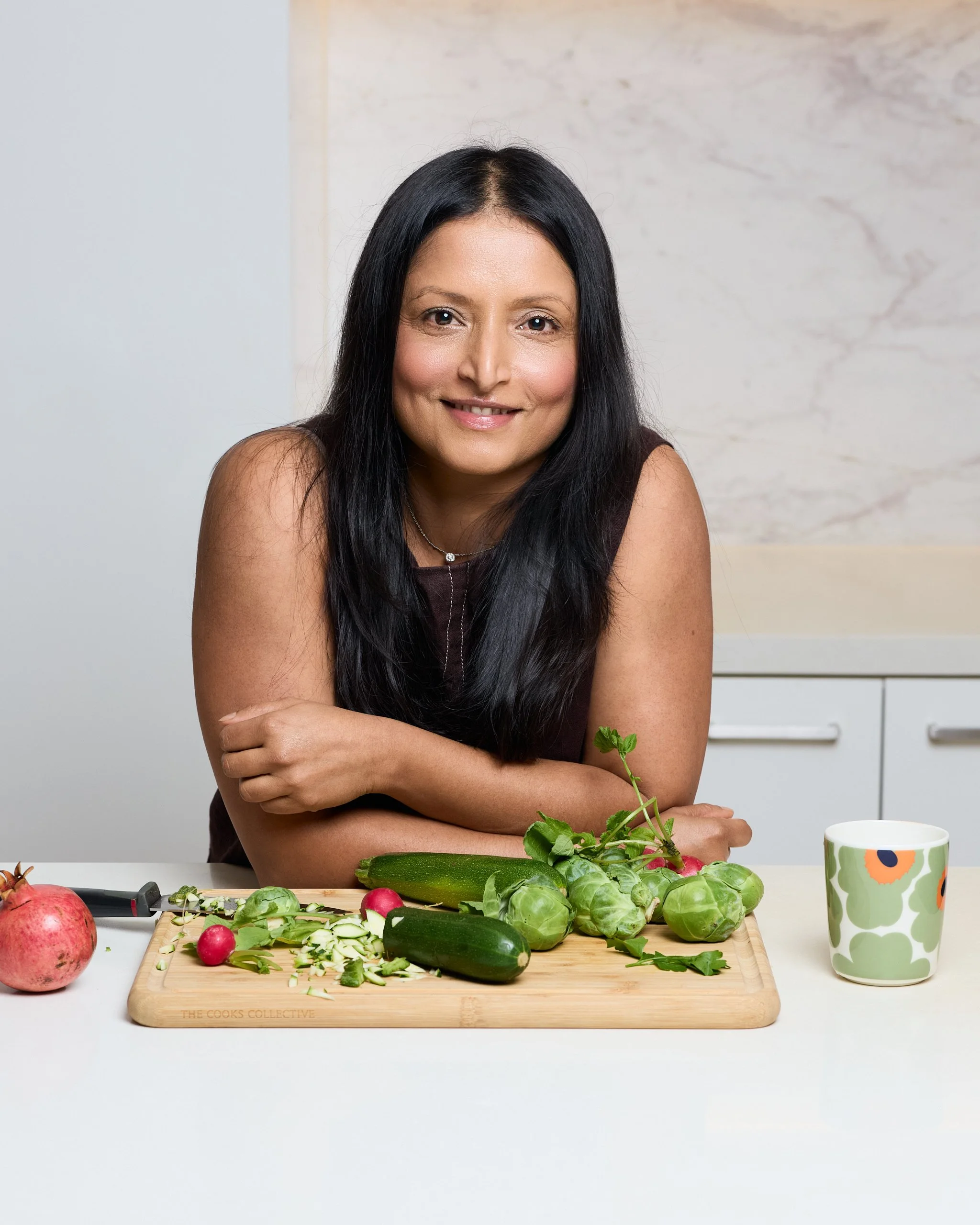 A woman with long black hair smiling at the camera while preparing vegetables on a wooden cutting board. There are radishes, cucumbers, and leafy greens on the board, with a pomegranate and a cucumber visible on the white countertop. The woman is in a modern kitchen with a white and marble background, and a ceramic mug with a floral pattern is on the right side of the image.