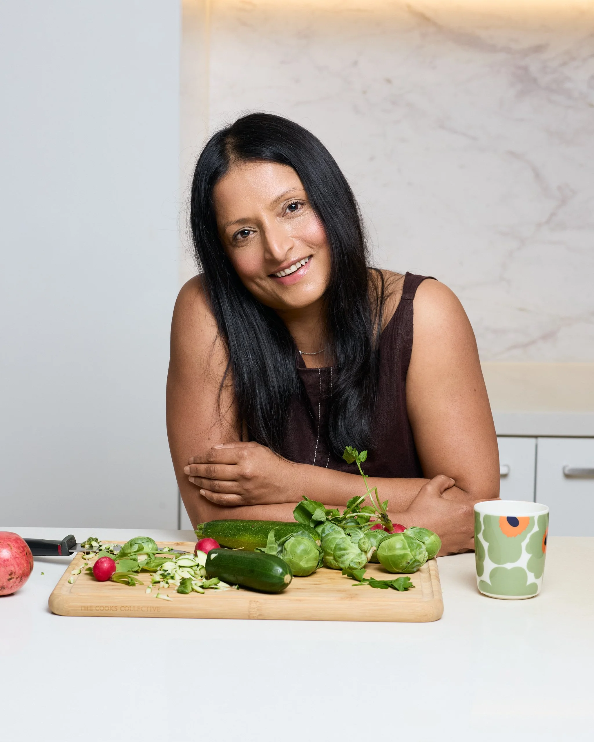 Woman with long black hair smiling at camera in a kitchen, surrounded by fresh vegetables including radishes, cucumbers, and leafy greens on a wooden cutting board, with a white and green patterned mug nearby.