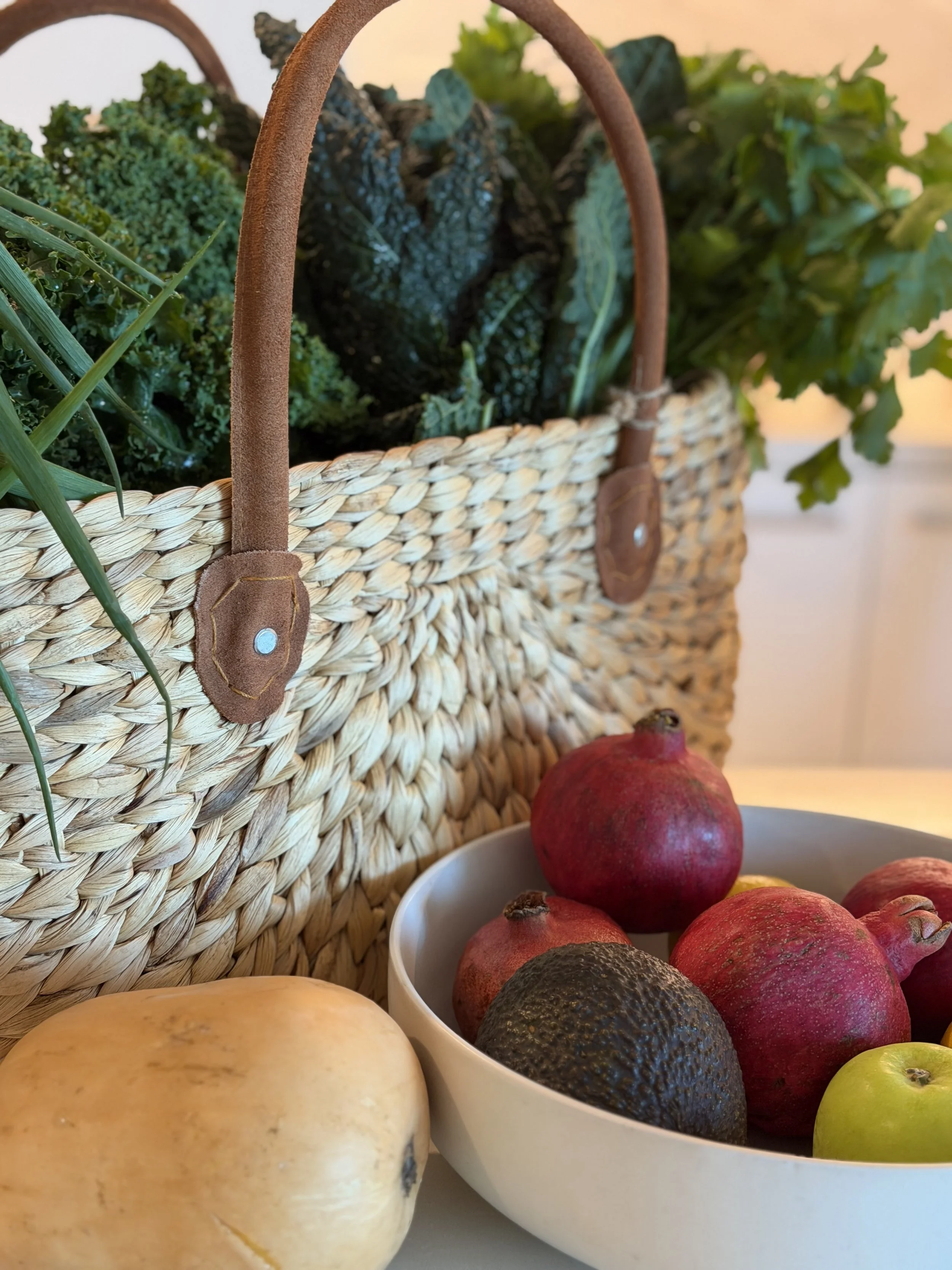 Close-up of a woven basket filled with leafy greens and purple radishes, with pomegranates, apples, and a melon in the foreground.