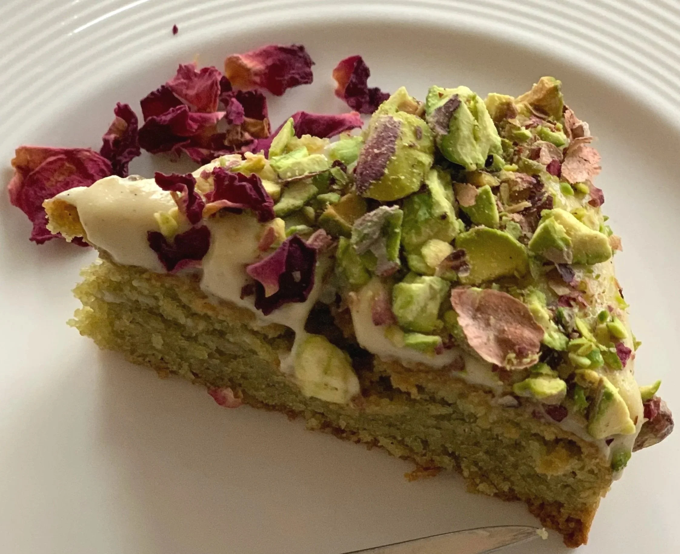 Close-up of a slice of cake topped with crushed pistachios and dried rose petals on a white dish.