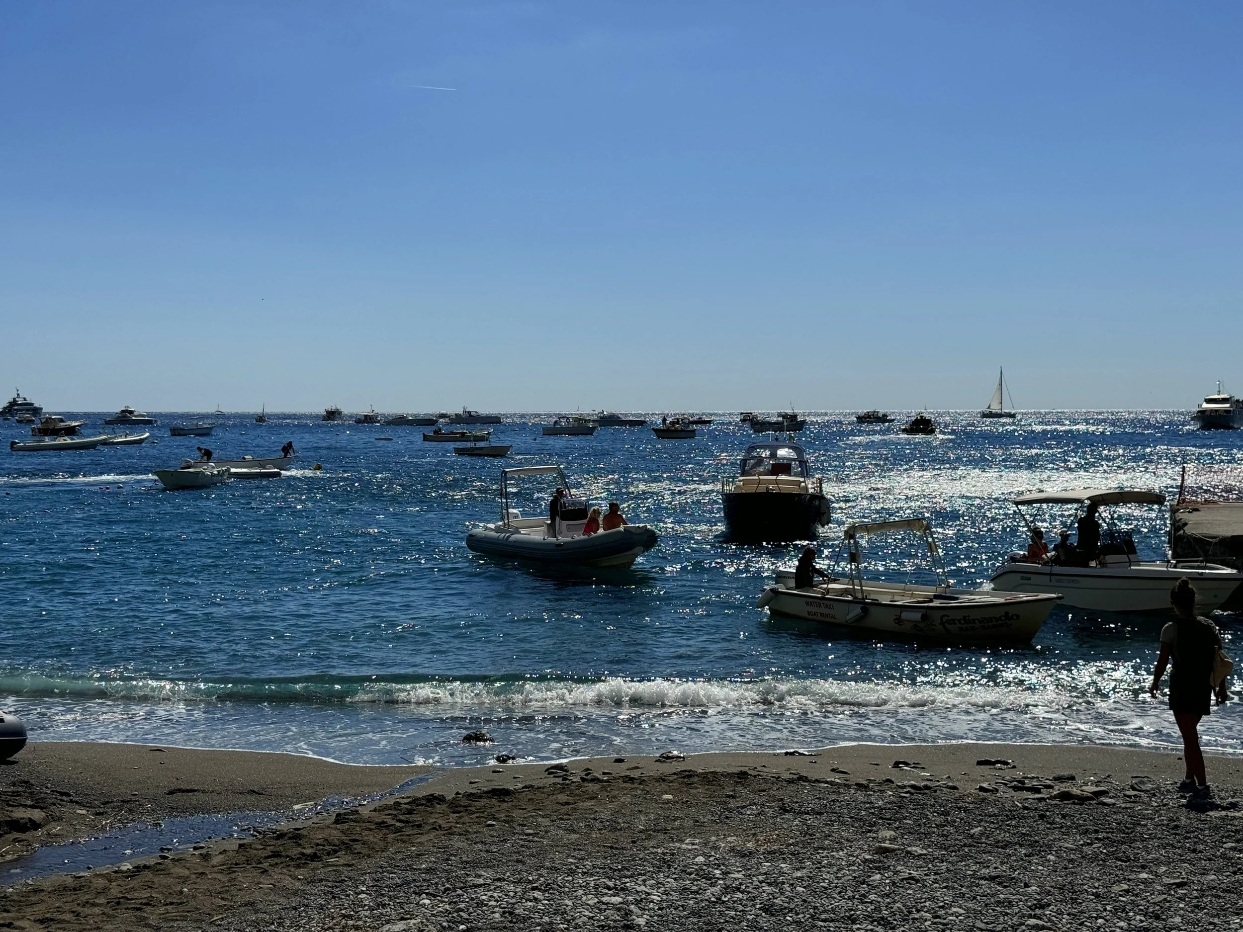 A beach scene with several boats anchored in the water, people walking along the shoreline, and a sailboat in the distance under a clear blue sky.