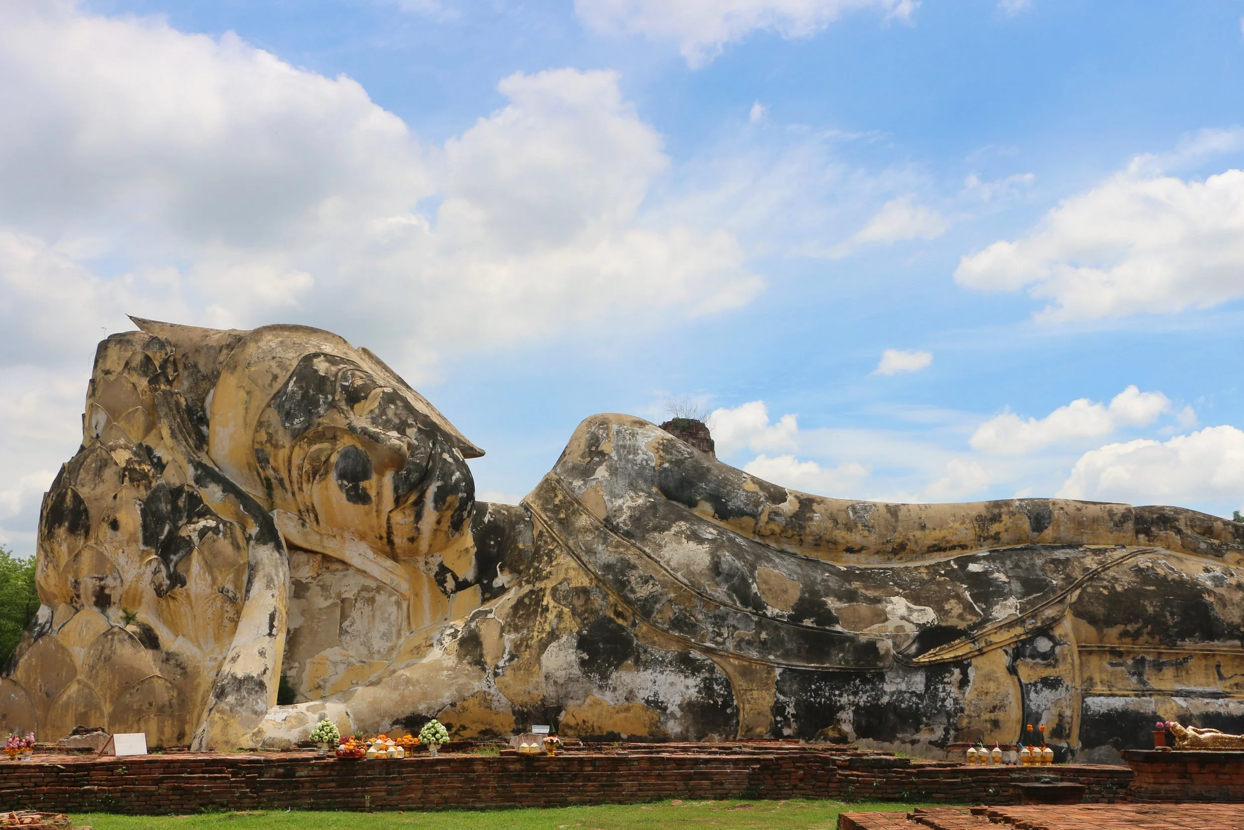 Large reclining Buddha statue made of stone with a weathered appearance, situated outdoors under a blue sky with scattered clouds. Small offerings and flowers are placed at the base of the statue.