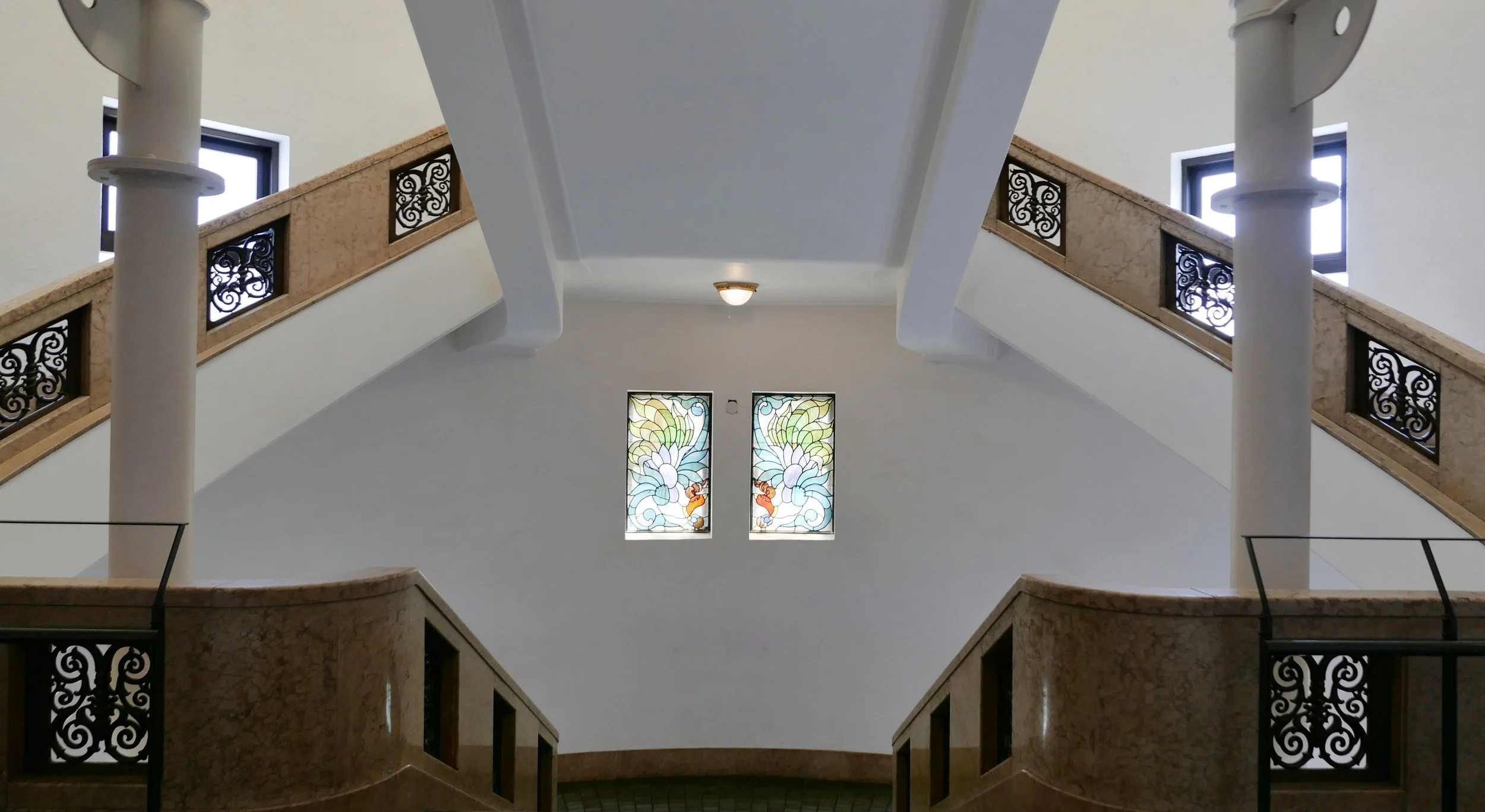 Interior view of a staircase with decorative wrought iron railings, marble steps, stained glass windows depicting colorful flowers, and two small windows near the ceiling allowing natural light to enter.