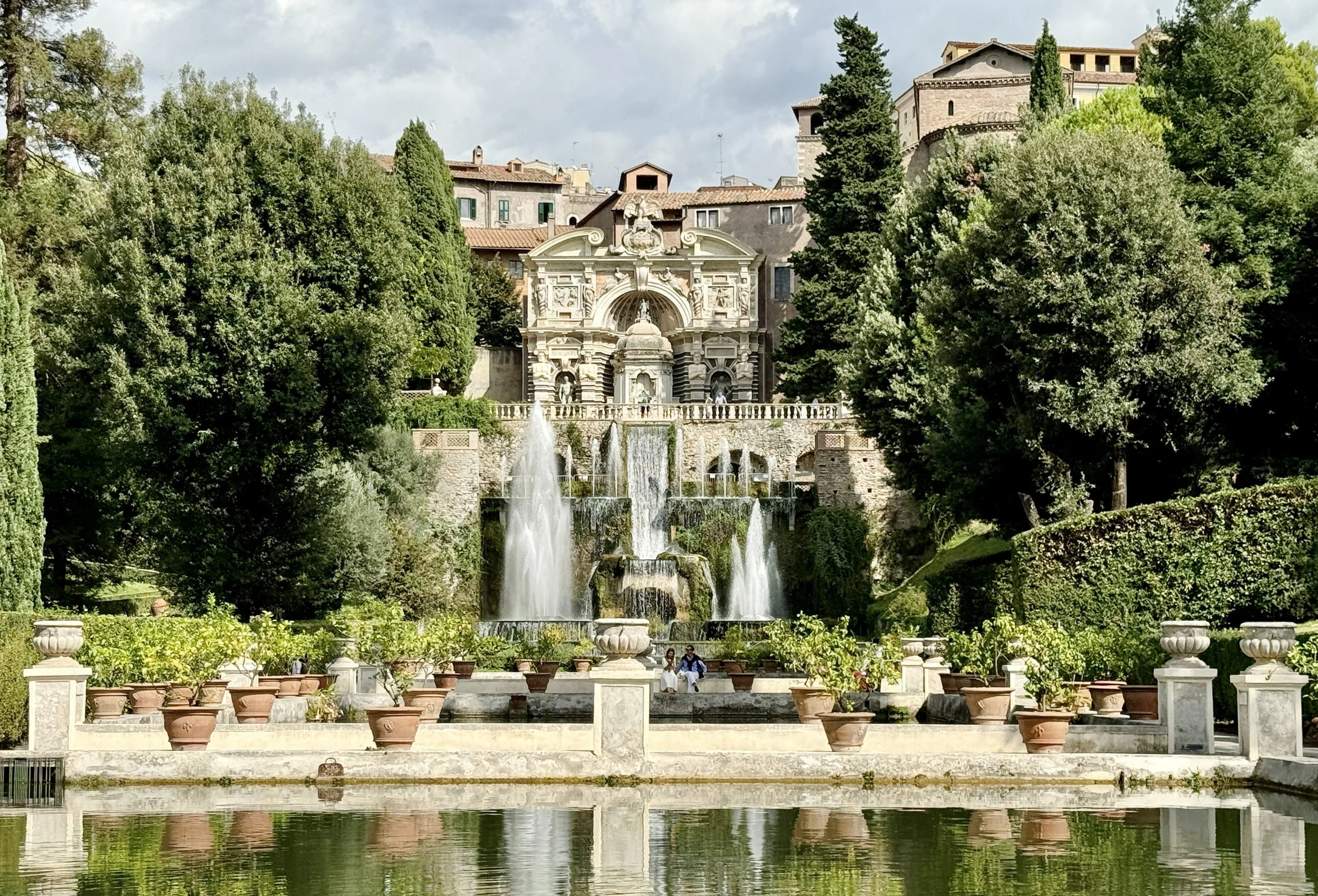 A scenic outdoor park with a fountain, trees, and historic building in the background, with people relaxing by the water.
