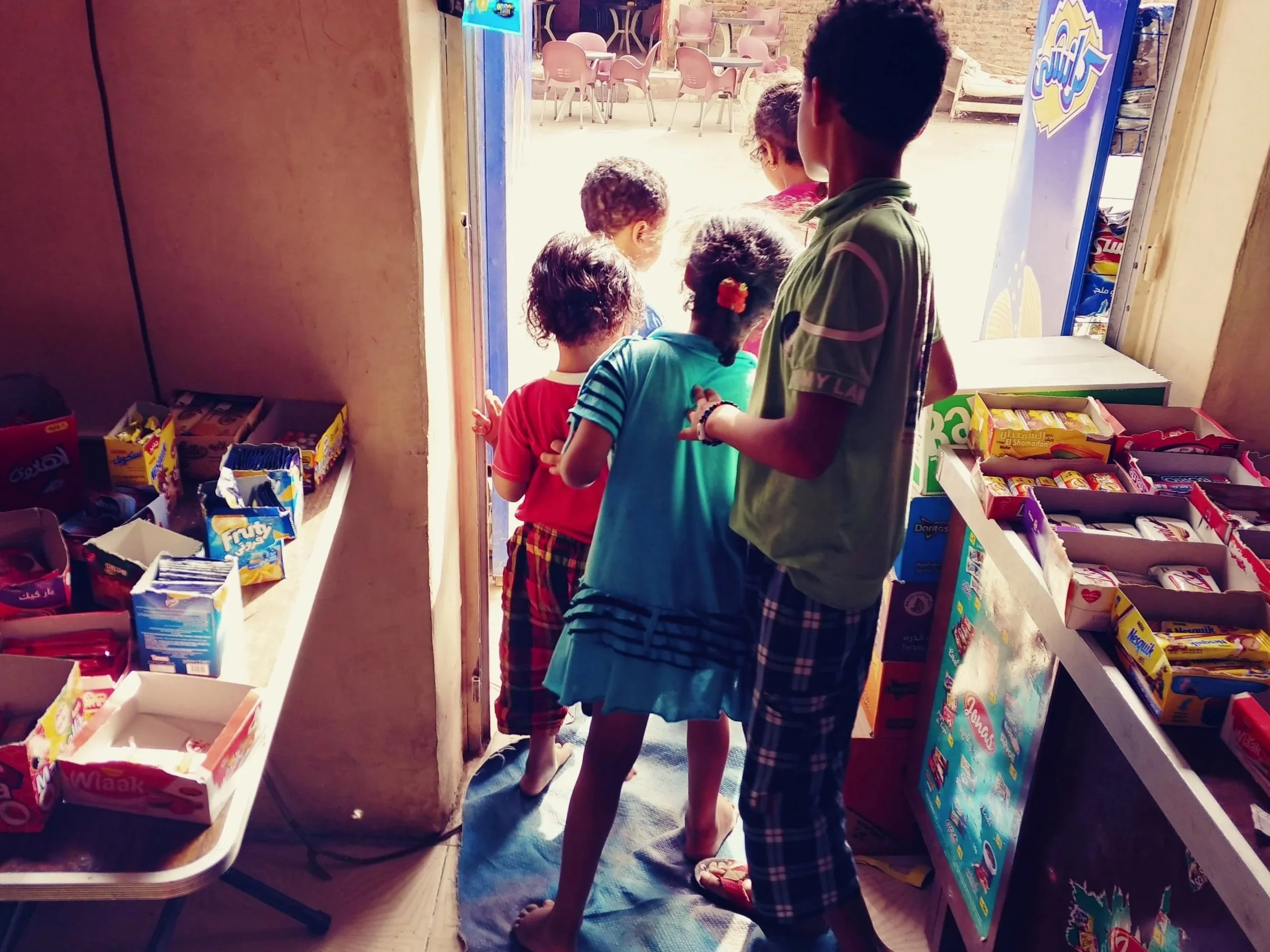 Children and an adult inside a store, looking at snack shelves near the entrance. Several children in colorful clothes are visible from the back, standing close to an adult, who is wearing a green and gray striped shirt. Bright sunlight is coming fro