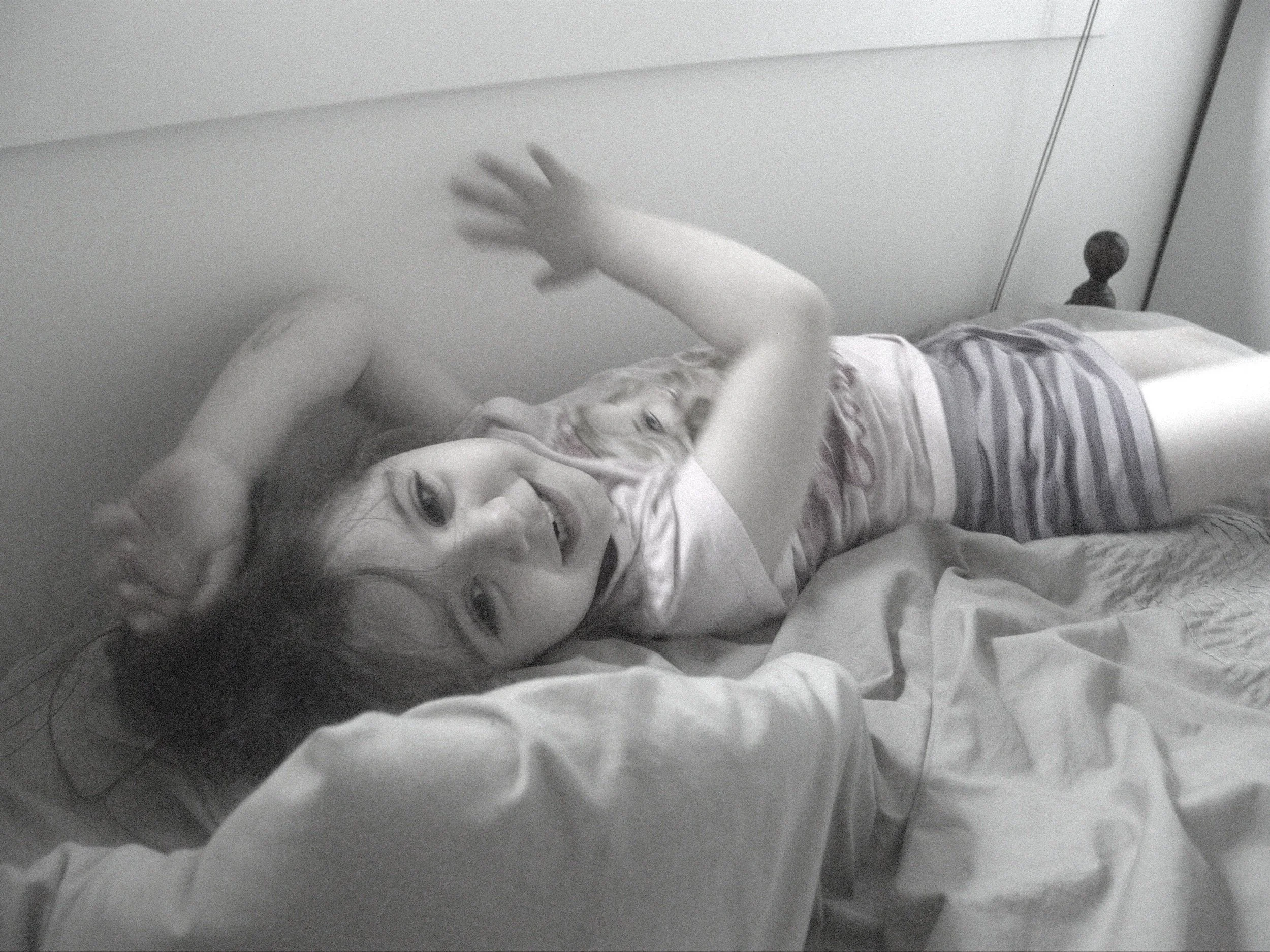 A young girl lying on a bed, smiling and reaching out with one hand, wearing striped pajamas, with her head resting on pillows.